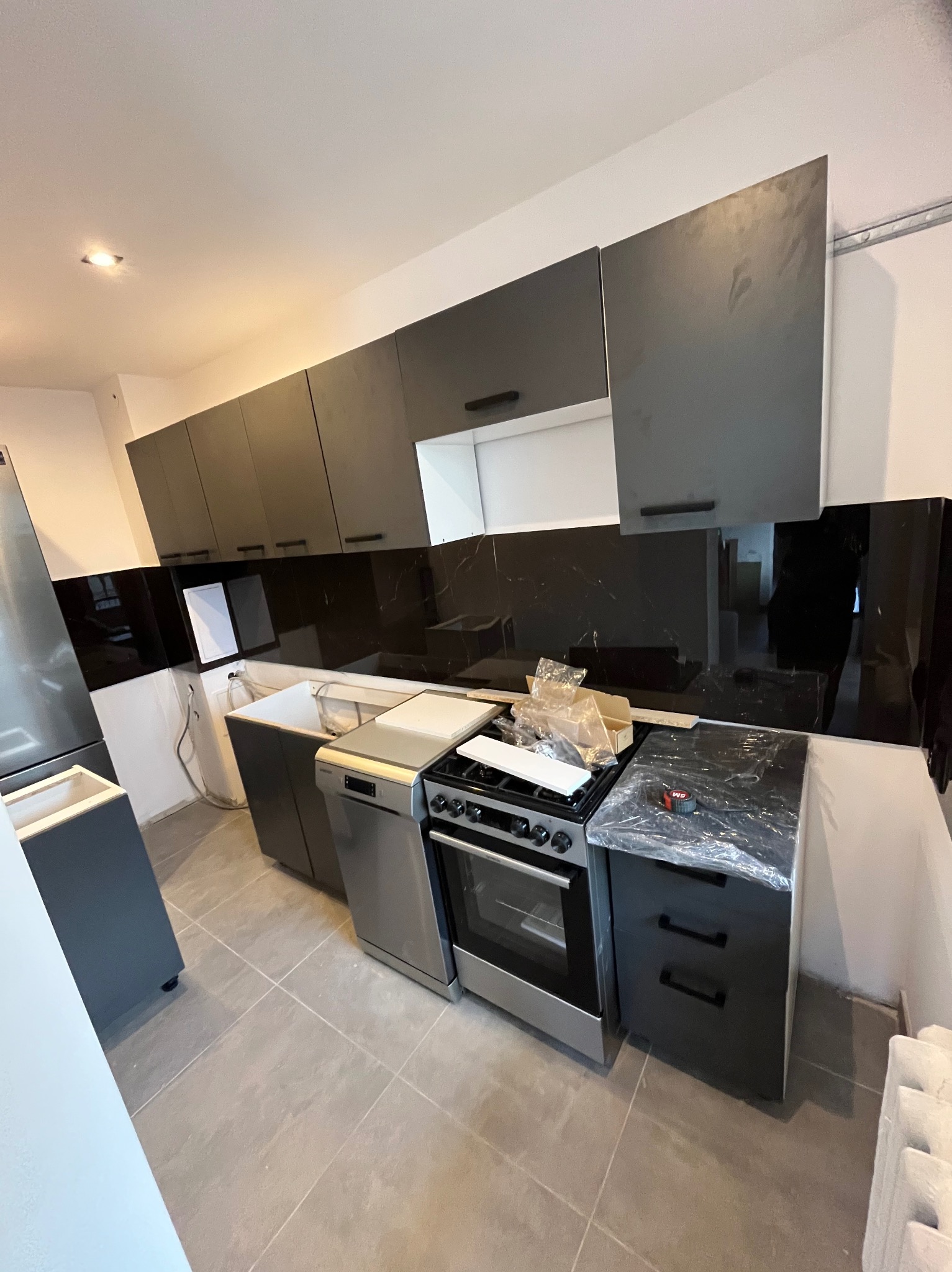 Nowoczesna kuchnia w trakcie montażu: szare szafki wiszące i dolne, czarny panel nad blatem, sprzęt AGD częściowo zabezpieczony folią. Widoczne niedokończone prace.