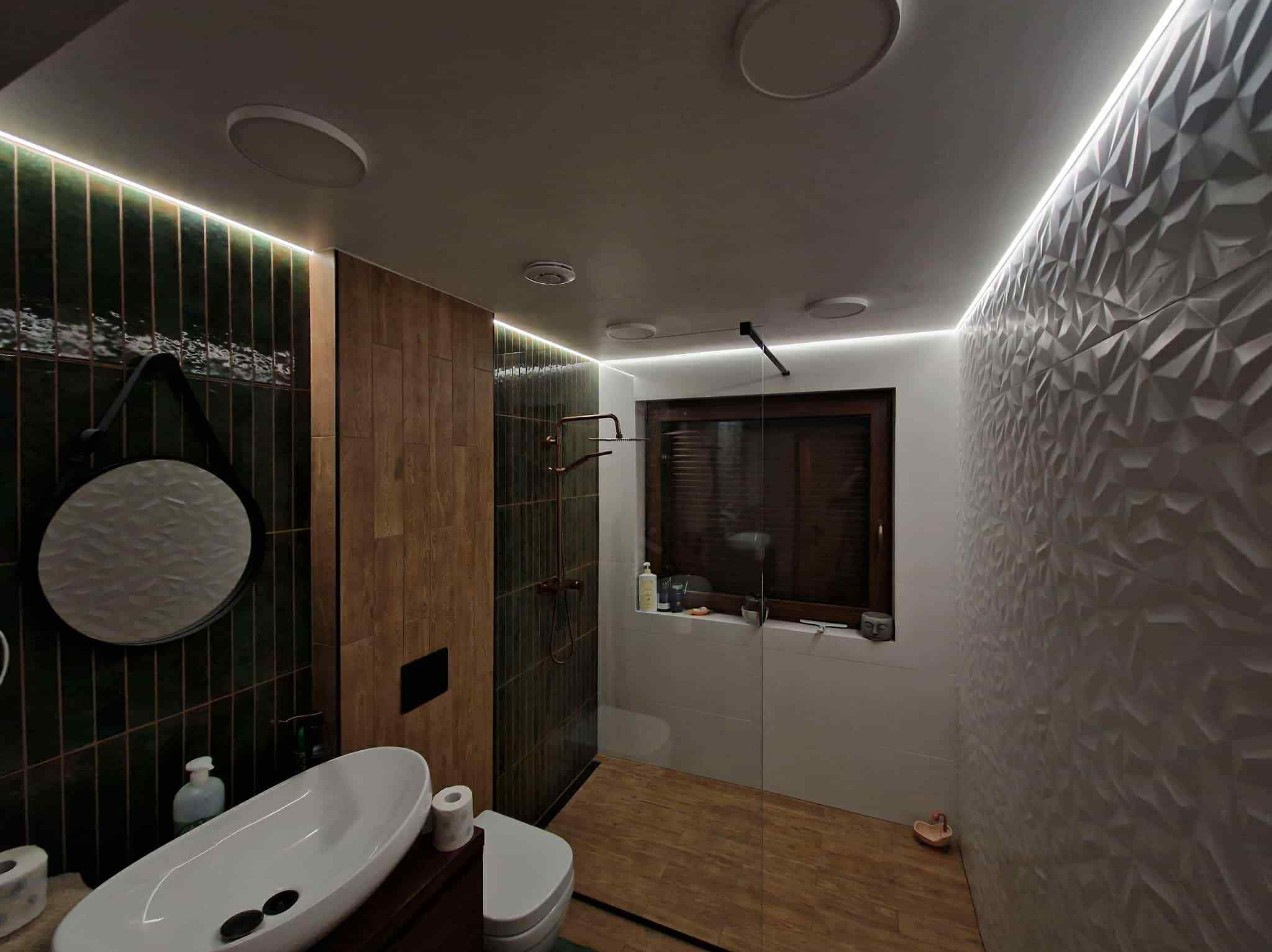 Nowoczesna łazienka z zielonymi i drewnianymi płytkami, kabiną prysznicową i umywalką na blacie. Oświetlenie LED podkreśla geometryczną fakturę ściany.