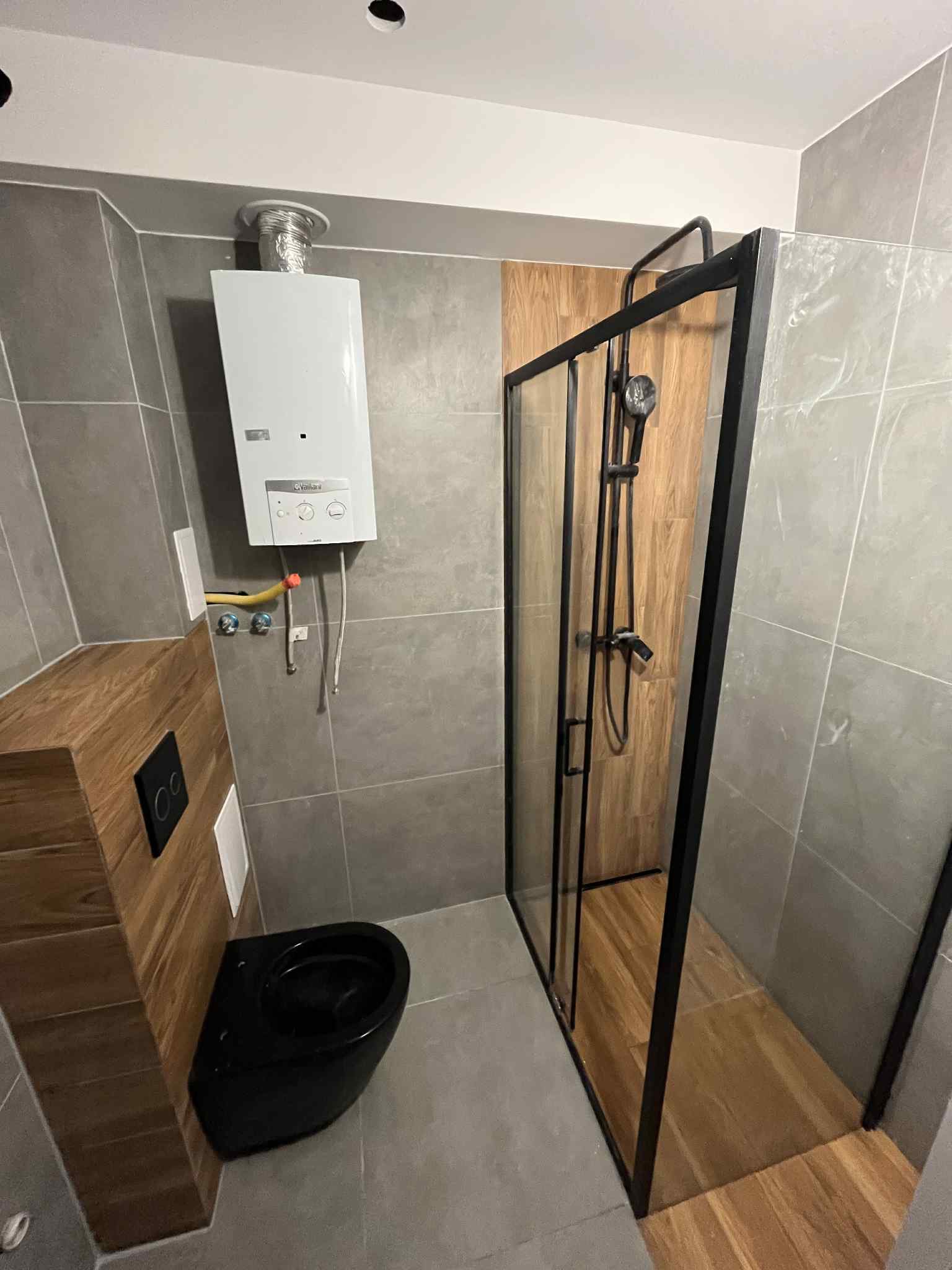 Wykończona łazienka z szarymi płytkami, czarną armaturą i kabiną prysznicową z czarnymi profilami. Widoczny piec gazowy i drewnopodobna obudowa toalety.