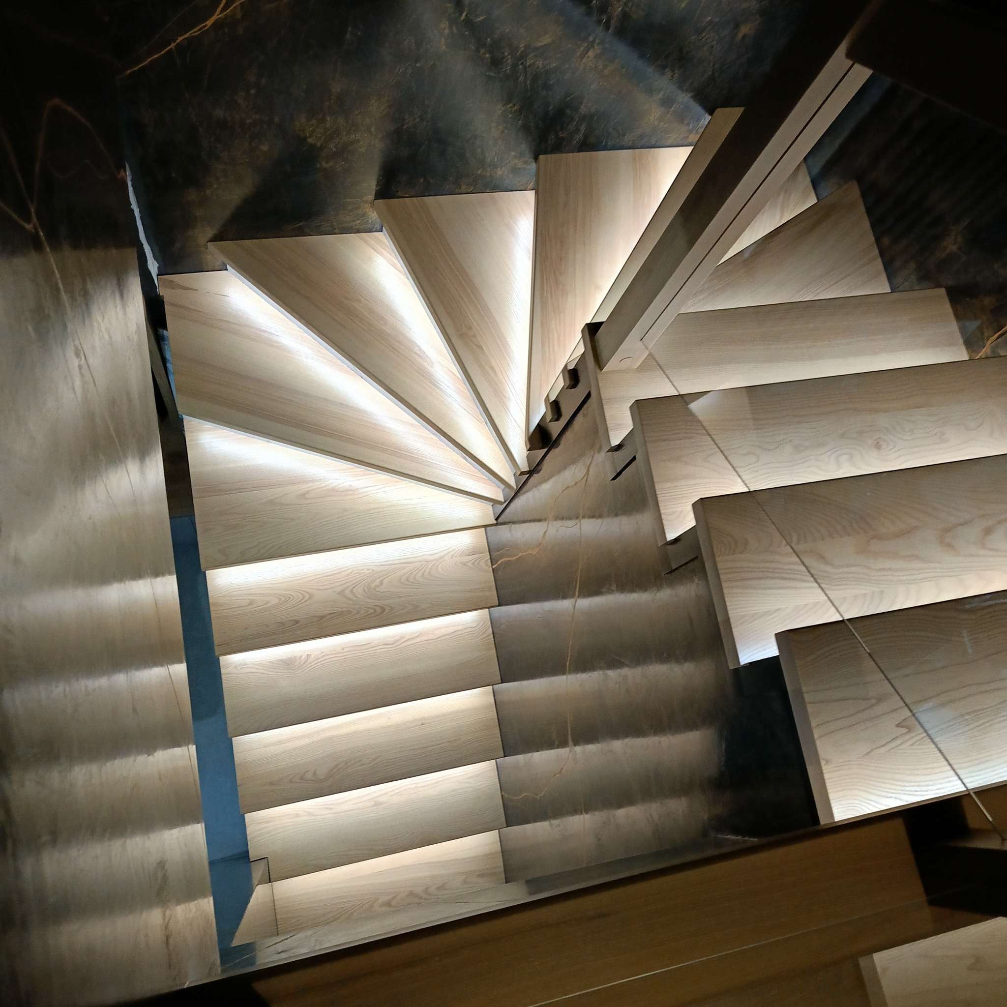 Spiralne schody z jasnego drewna z subtelnym oświetleniem LED pod każdym stopniem, widziane z góry. Ciemne tło ściany podkreśla nowoczesny design.