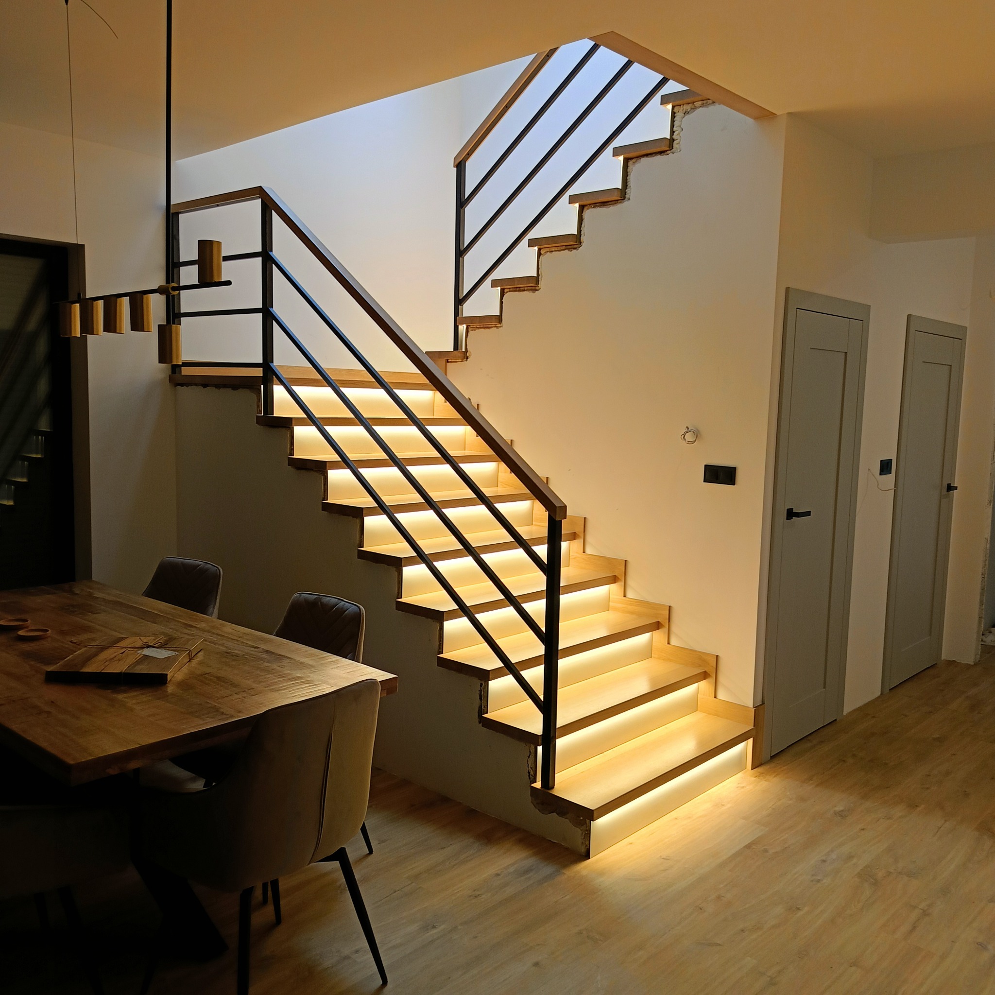 Nowoczesne schody drewniane z podświetleniem LED, czarna balustrada. Widok z dołu na jasne stopnie i minimalistyczny design. W tle stół i krzesła.