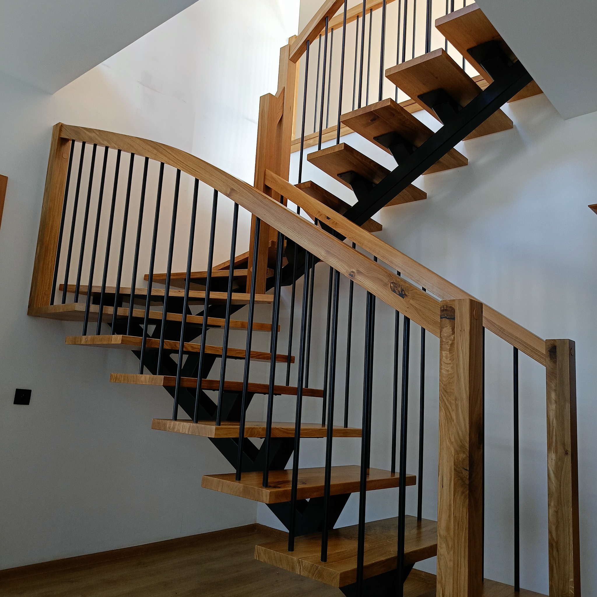 Drewniane schody z czarnymi metalowymi elementami w nowoczesnym wnętrzu. Jasne drewno stopni kontrastuje z ciemnymi balustradami i wspornikami.