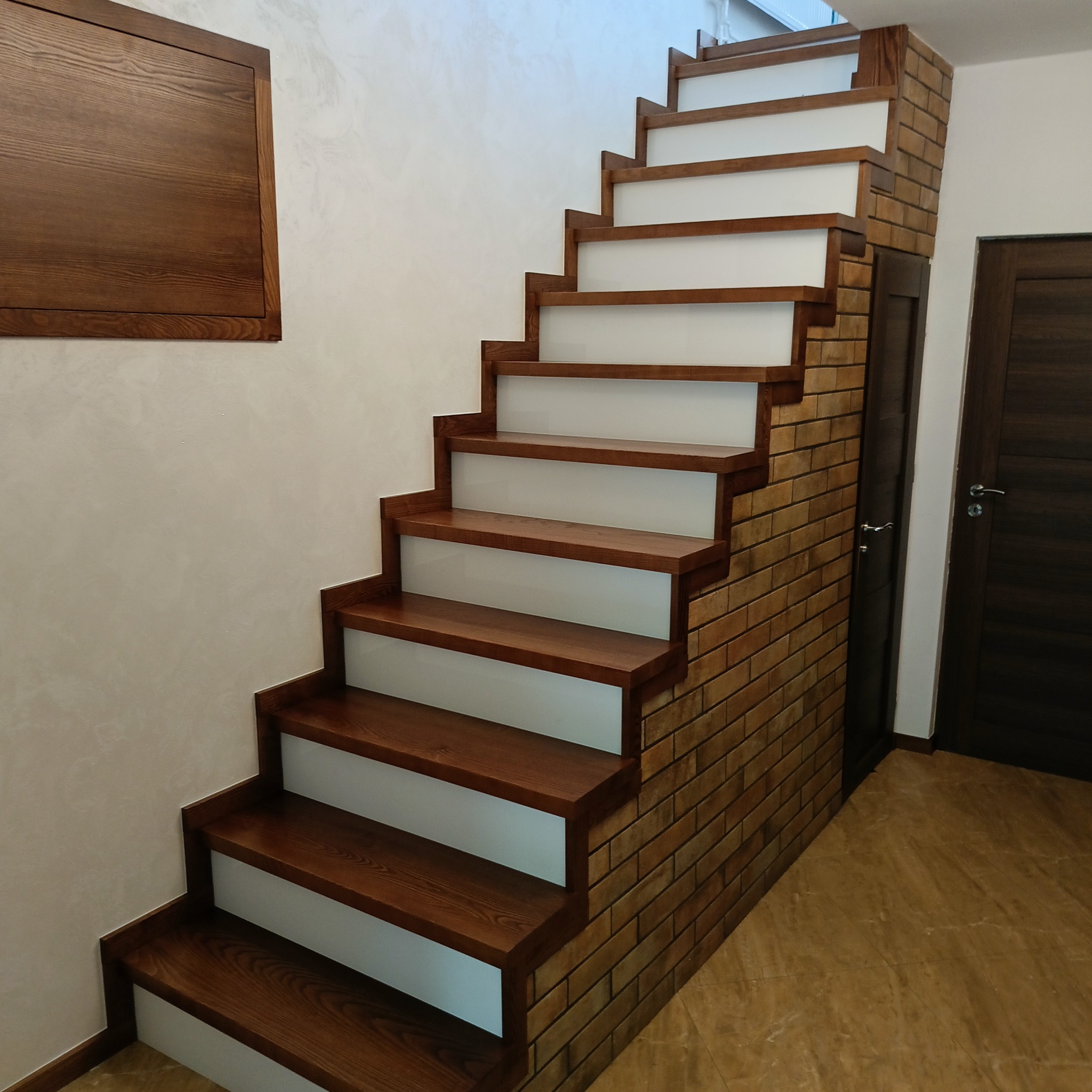 Drewniane schody z białymi podstopnicami, wykończone cegłą przy ścianie, obok ciemne drzwi. Na ścianie dekoracyjny panel z drewna.