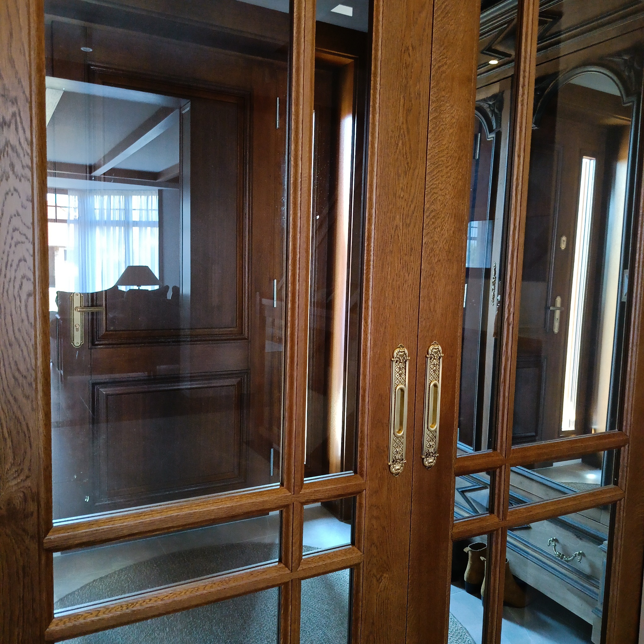 Szklane drzwi wewnętrzne z drewnianą ramą i ozdobnymi, złotymi klamkami. Widoczne odbicie wnętrza domu. Ciemne drewno i klasyczny styl.