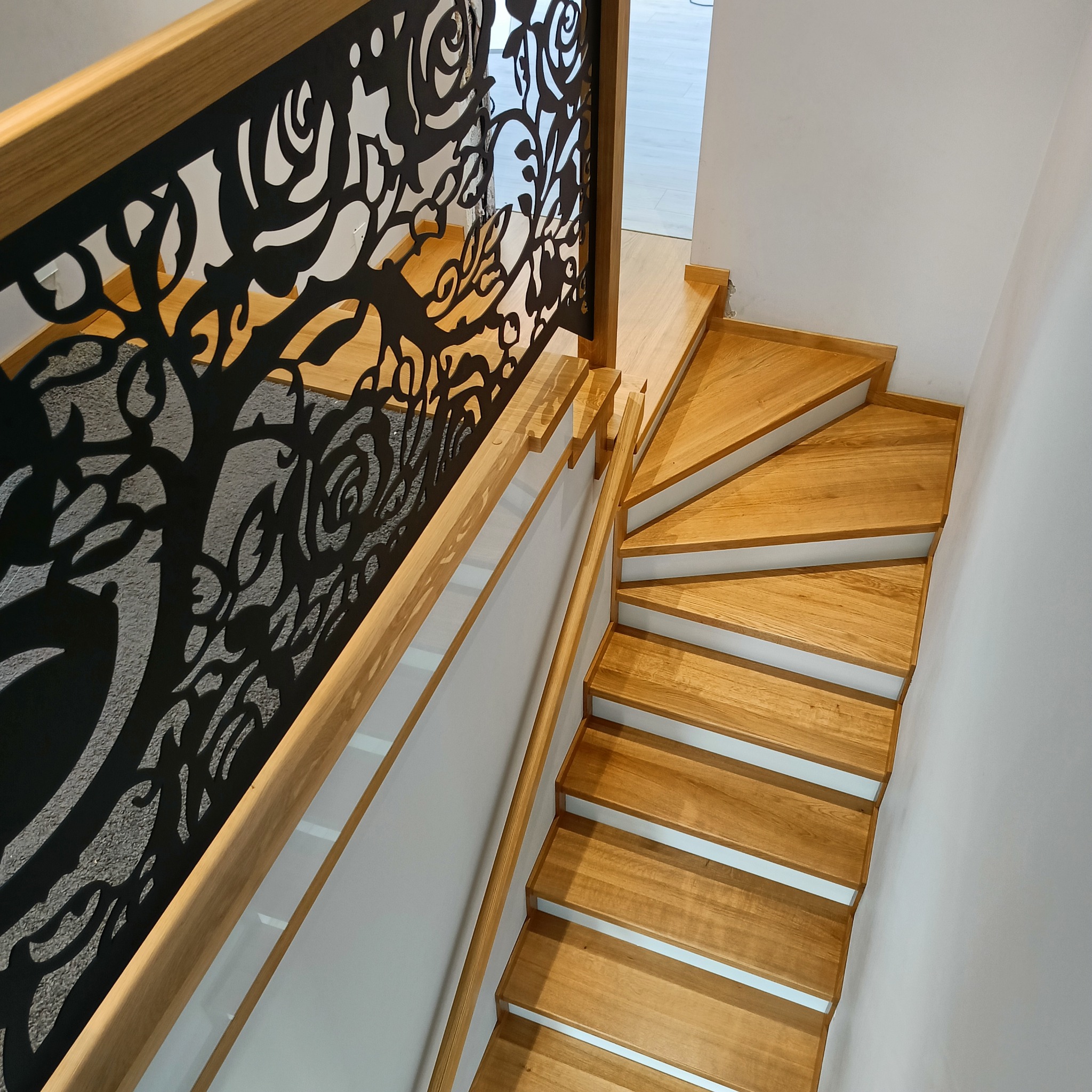 Nowoczesne schody drewniane z ażurową balustradą o motywie roślinnym, prowadzące na wyższą kondygnację. Jasne drewno, białe ściany, minimalistyczny design.