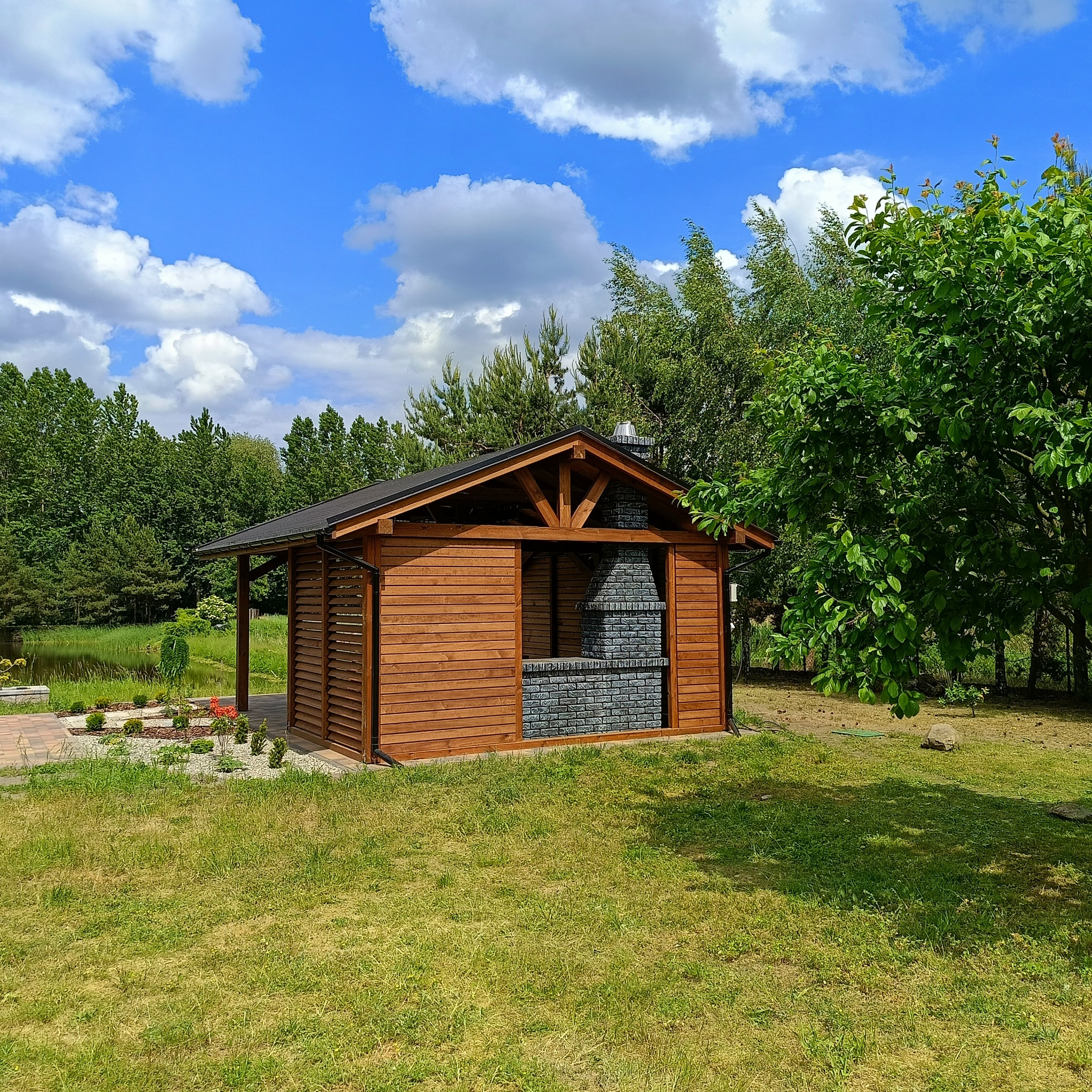 Drewniana altana z grillem z szarej cegły na tle zieleni i błękitnego nieba. Konstrukcja zadaszona, z elementami dekoracyjnymi. Otoczenie: trawnik, drzewa.