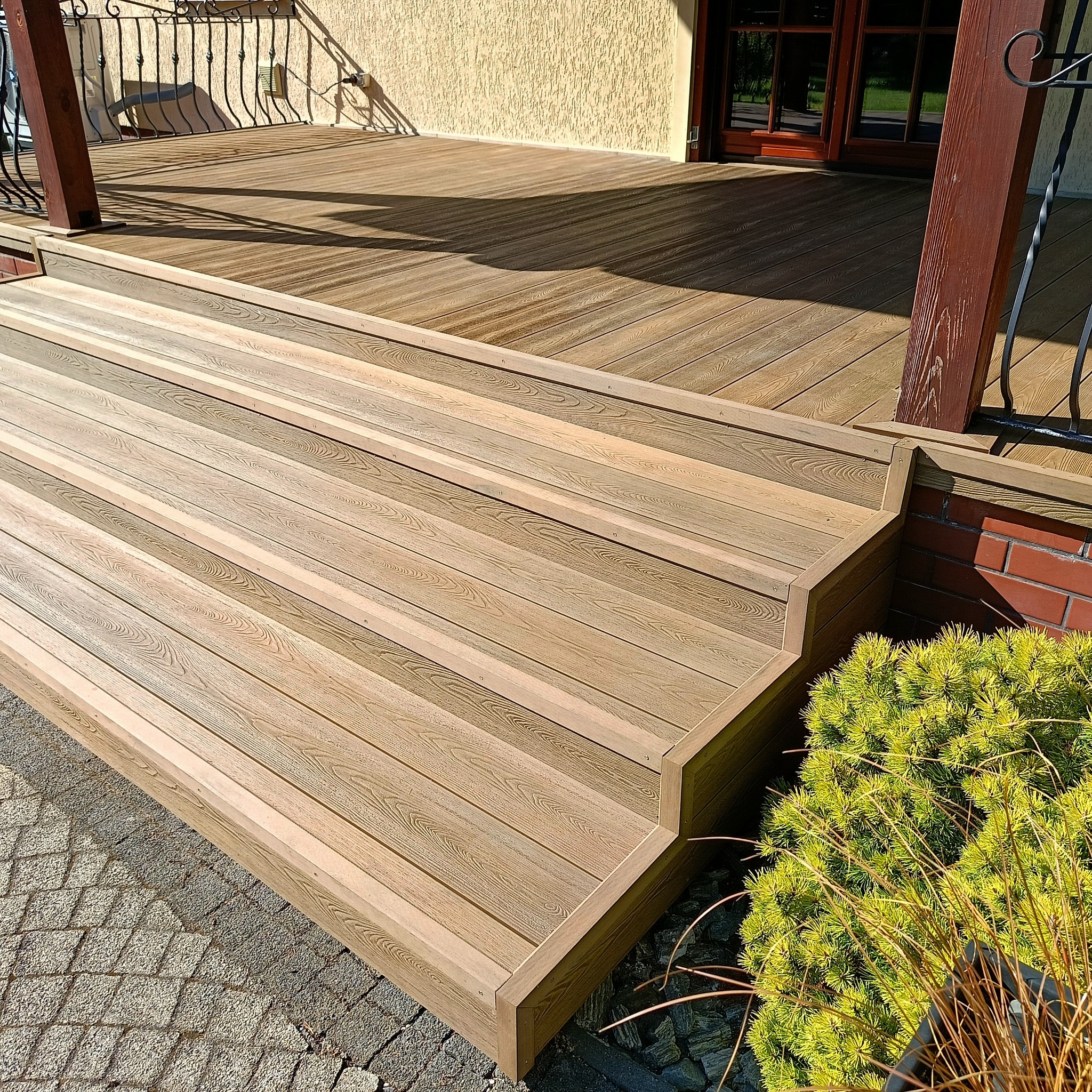 Nowy taras z kompozytu drewna z niskimi schodami i dekoracyjną roślinnością. Widok na dom z beżową elewacją i ciemnymi oknami.