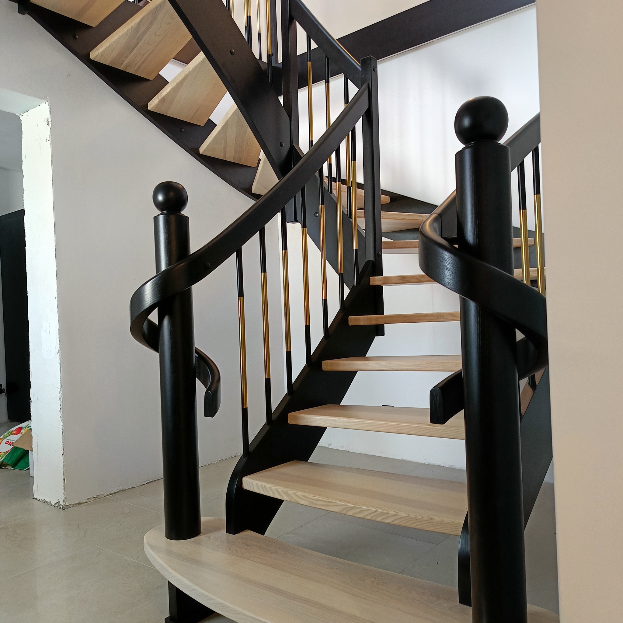 Nowoczesne schody drewniane z czarną balustradą i złotymi elementami. Jasne stopnie kontrastują z ciemną konstrukcją. Widoczne detale wykończenia.