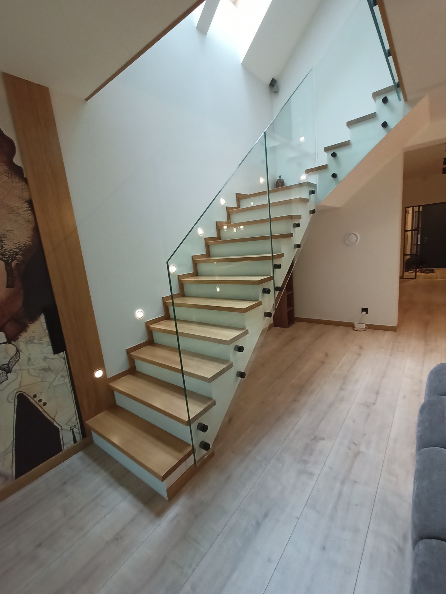 Nowoczesne schody drewniane z podświetleniem LED i szklaną balustradą, widok z dołu, jasne drewno, minimalistyczny design, oświetlenie punktowe.