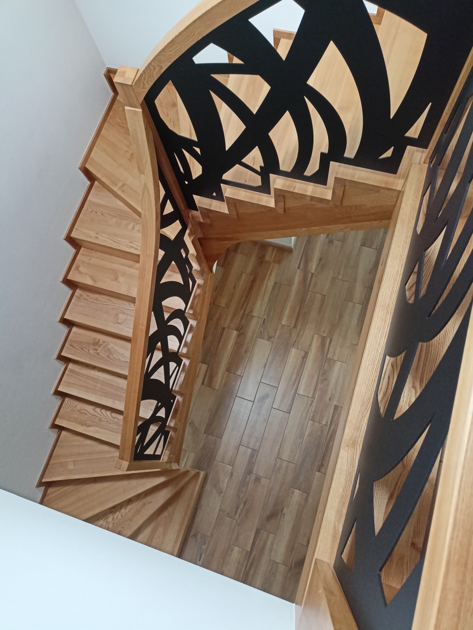 Kątowe ujęcie z góry na drewniane schody spiralne z ozdobną, czarną balustradą. Widoczne drewniane stopnie i podłoga z płytek imitujących drewno.