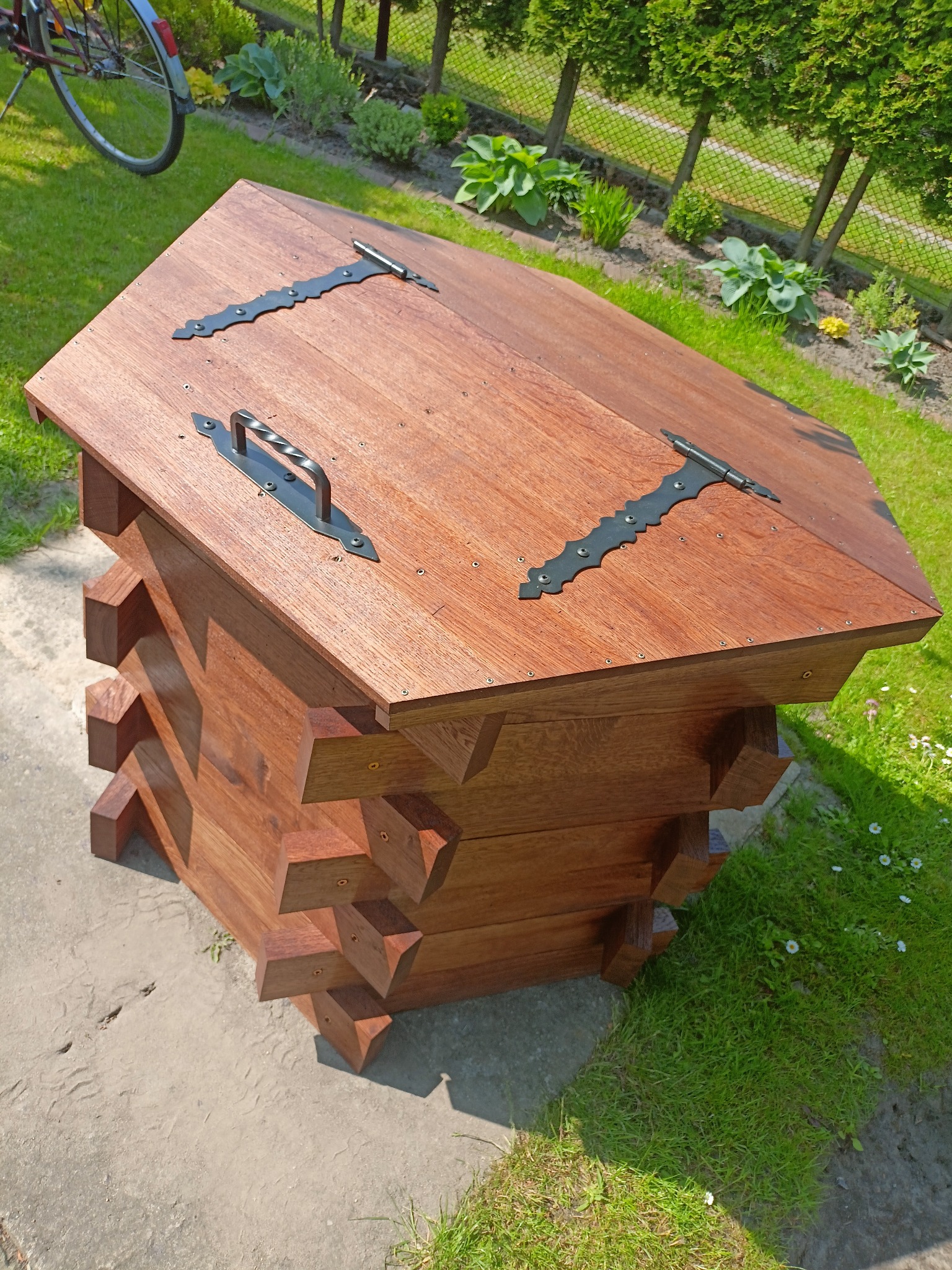 Drewniana skrzynia ogrodowa z pokrywą, zdobiona metalowymi okuciami i uchwytem, w rustykalnym stylu, na tle zielonego trawnika i żywopłotu.