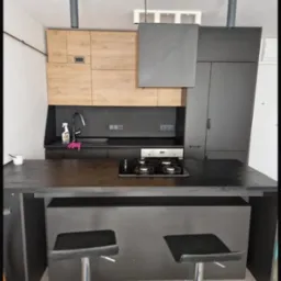 Nowoczesna kuchnia z wyspą: czarne fronty, drewniane akcenty, minimalistyczne lampy. Butelka środka czyszczącego na blacie sugeruje świeżość.