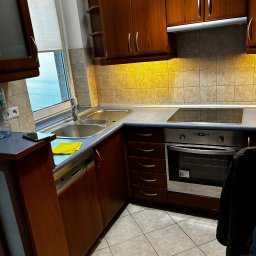 Instant Clean - Umyta kuchnia z drewnianymi szafkami, czystym zlewem i blatem. Na blacie leży żółta ściereczka. Widoczny piekarnik i płyta indukcyjna. Płytki na ścianie w odcieniach beżu.