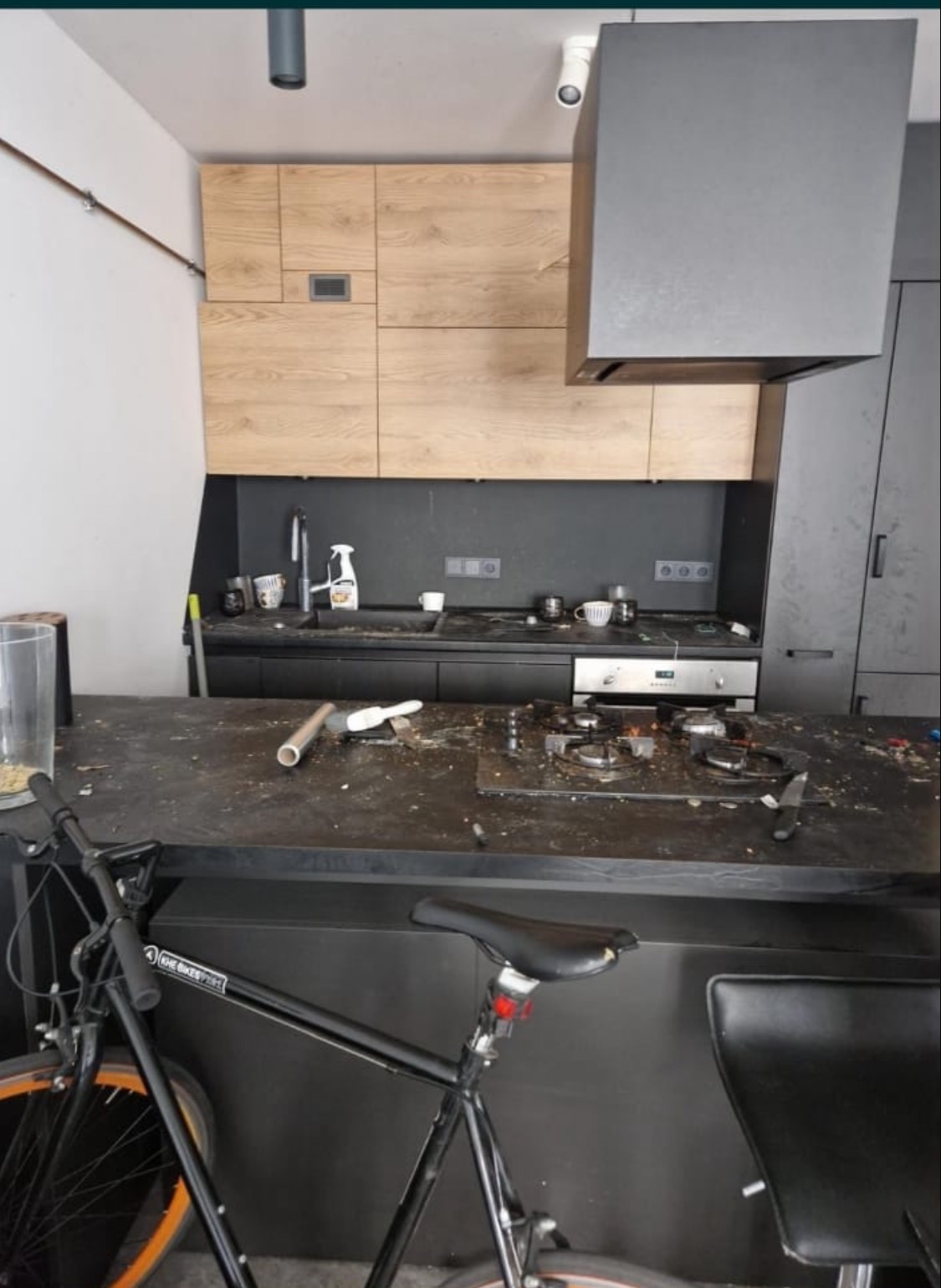 Zabrudzona kuchnia z czarnym blatem, widoczne resztki jedzenia i rower w tle. Ujęcie z góry na zanieczyszczony blat kuchenny, z elementami wyposażenia i rowerem.