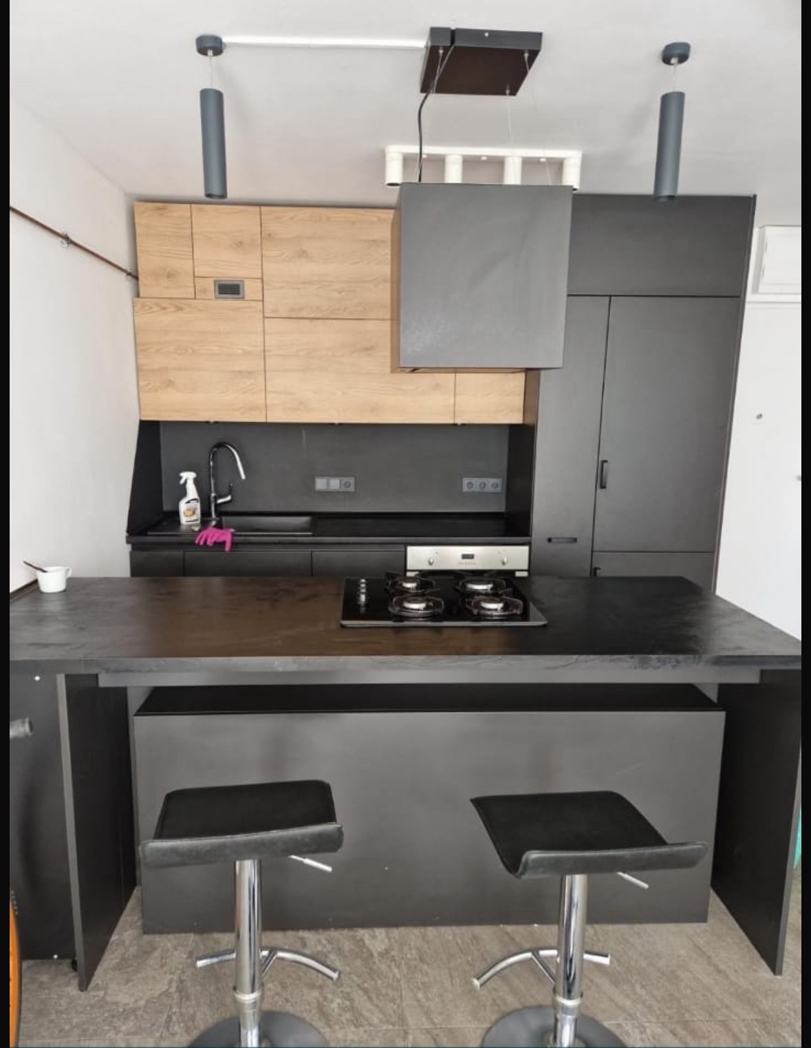 Nowoczesna kuchnia z wyspą: czarne fronty, drewniane akcenty, minimalistyczne lampy. Butelka środka czyszczącego na blacie sugeruje świeżość.