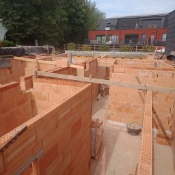 Roman Ząbczyk - Wznoszenie ścian działowych z czerwonej cegły ceramicznej, widok z góry na wnętrze budynku w trakcie budowy, drewniane podpory i narzędzia budowlane w kadrze.