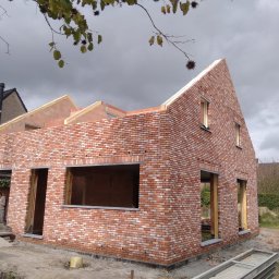 Roman Ząbczyk - Surowy budynek z czerwonej cegły w trakcie budowy, widoczne otwory okienne i drzwiowe, częściowo wykończone ściany, drabina budowlana z boku.