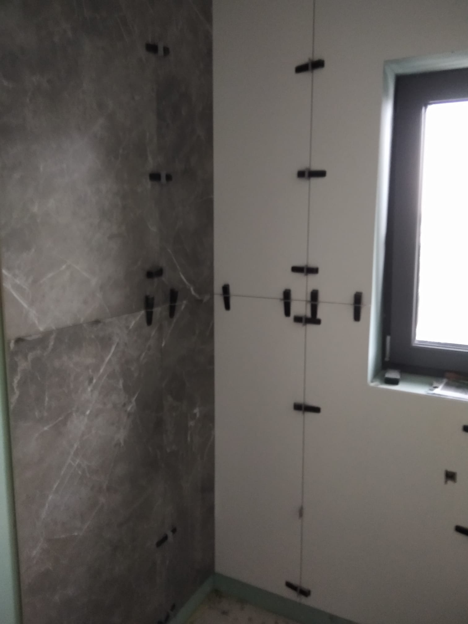 Ściana łazienki w trakcie wykańczania z szarymi płytkami imitującymi marmur i systemem poziomowania płytek, widoczne okno.