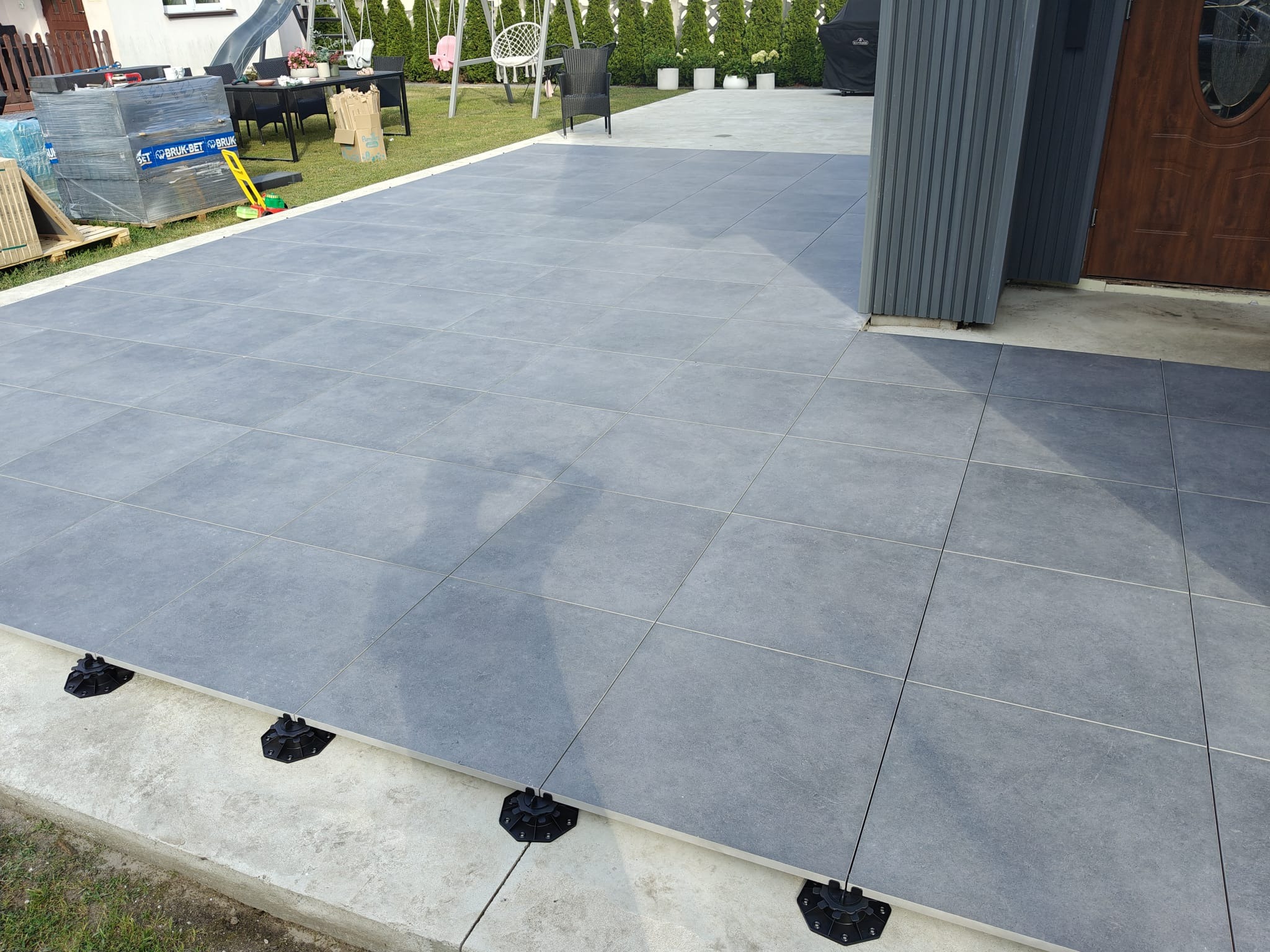 Szare płytki tarasowe na regulowanych wspornikach, widok z góry. Na trawniku w tle meble ogrodowe i zjeżdżalnia. Część płytek jeszcze nieułożona.