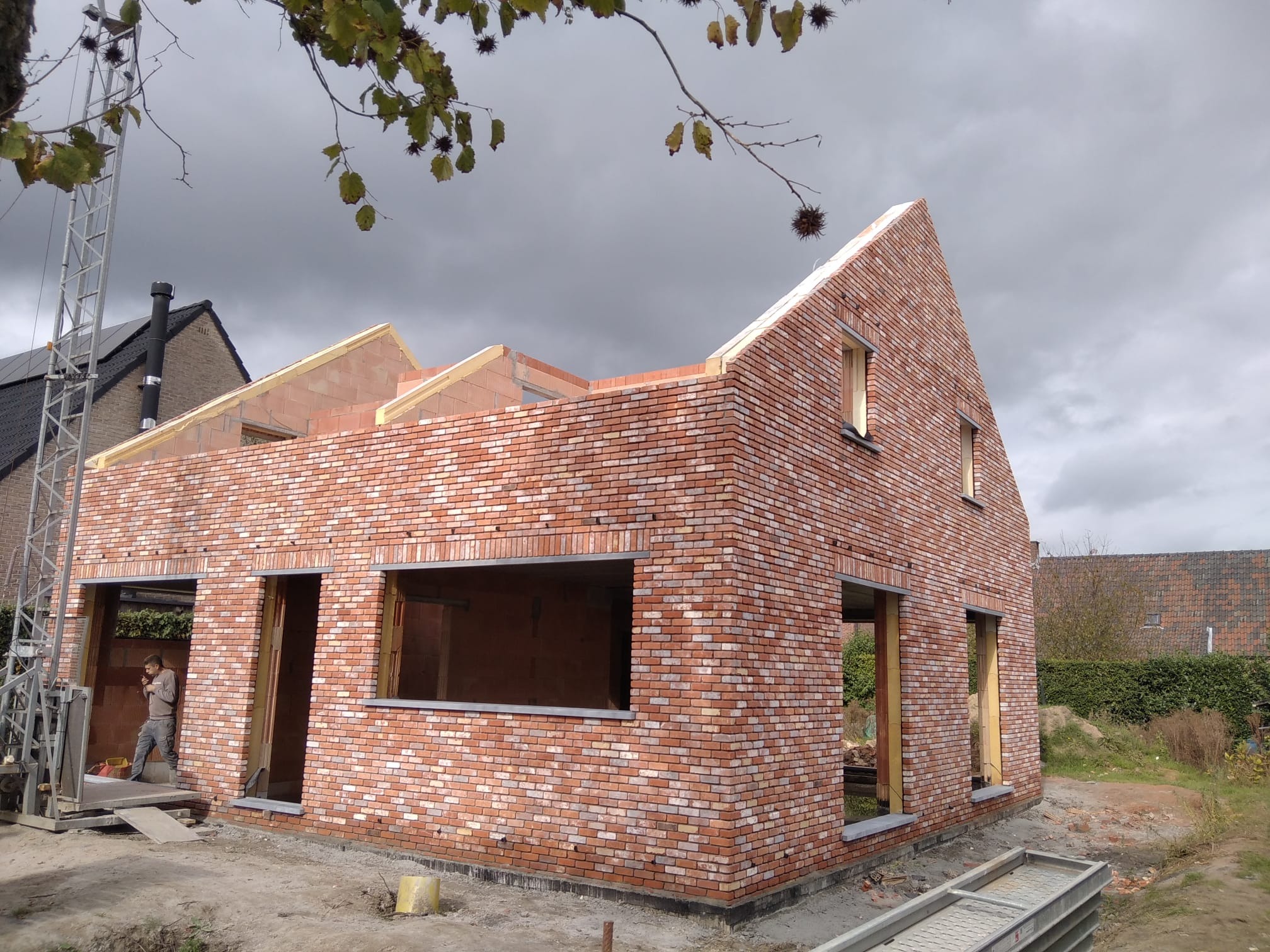 Surowy budynek z czerwonej cegły w trakcie budowy, widoczne otwory okienne i drzwiowe, częściowo wykończone ściany, drabina budowlana z boku.