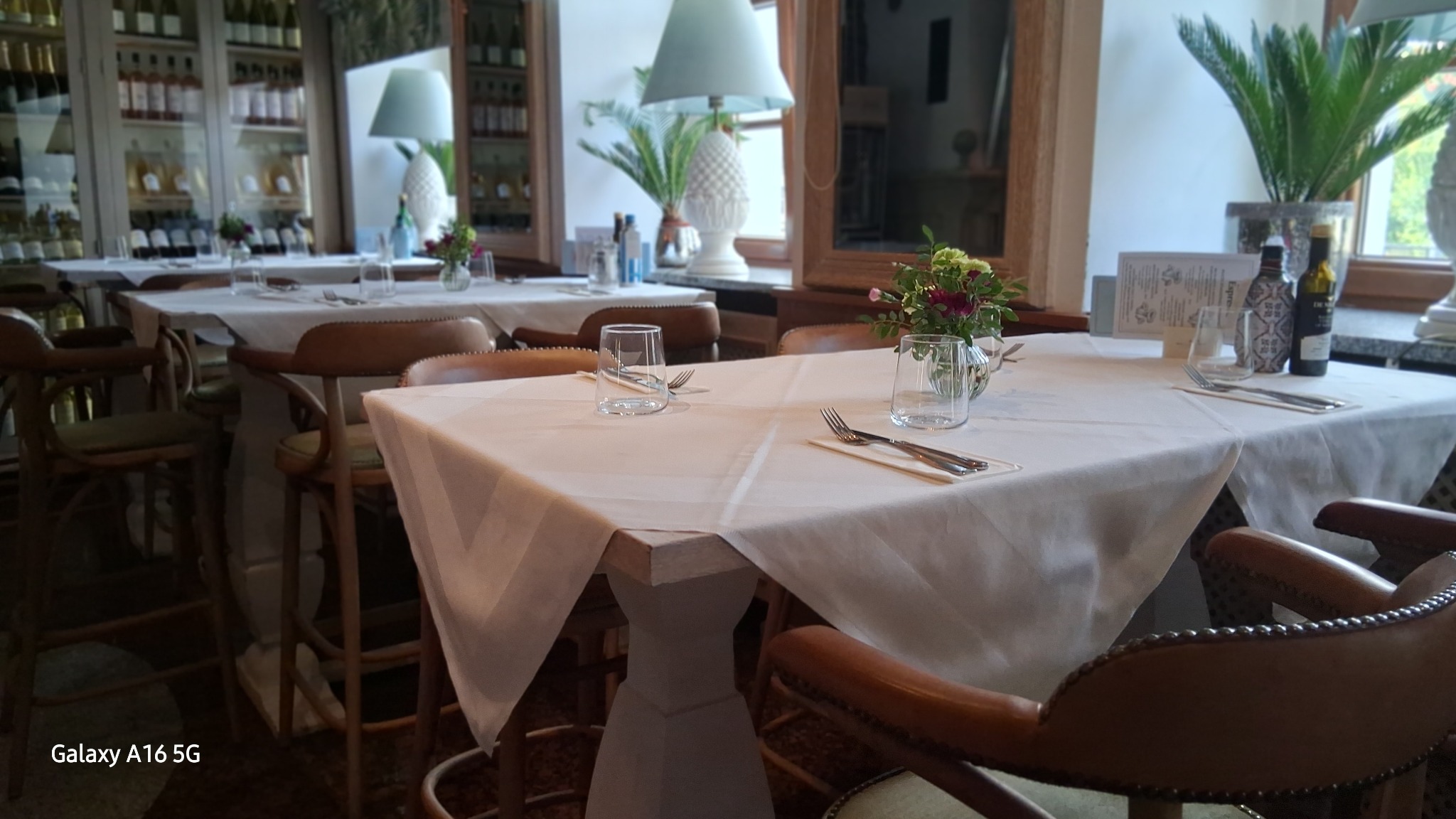 Elegancko nakryty stół w restauracji z białym obrusem, szklankami, sztućcami i kwiatami. W tle regał z butelkami i rośliny doniczkowe.