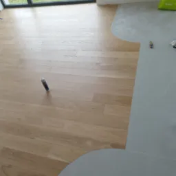 Połączenie paneli podłogowych z szarą posadzką. Widoczna tuba kleju, nóż i torba na tle okna. Układanie paneli w trakcie prac wykończeniowych.