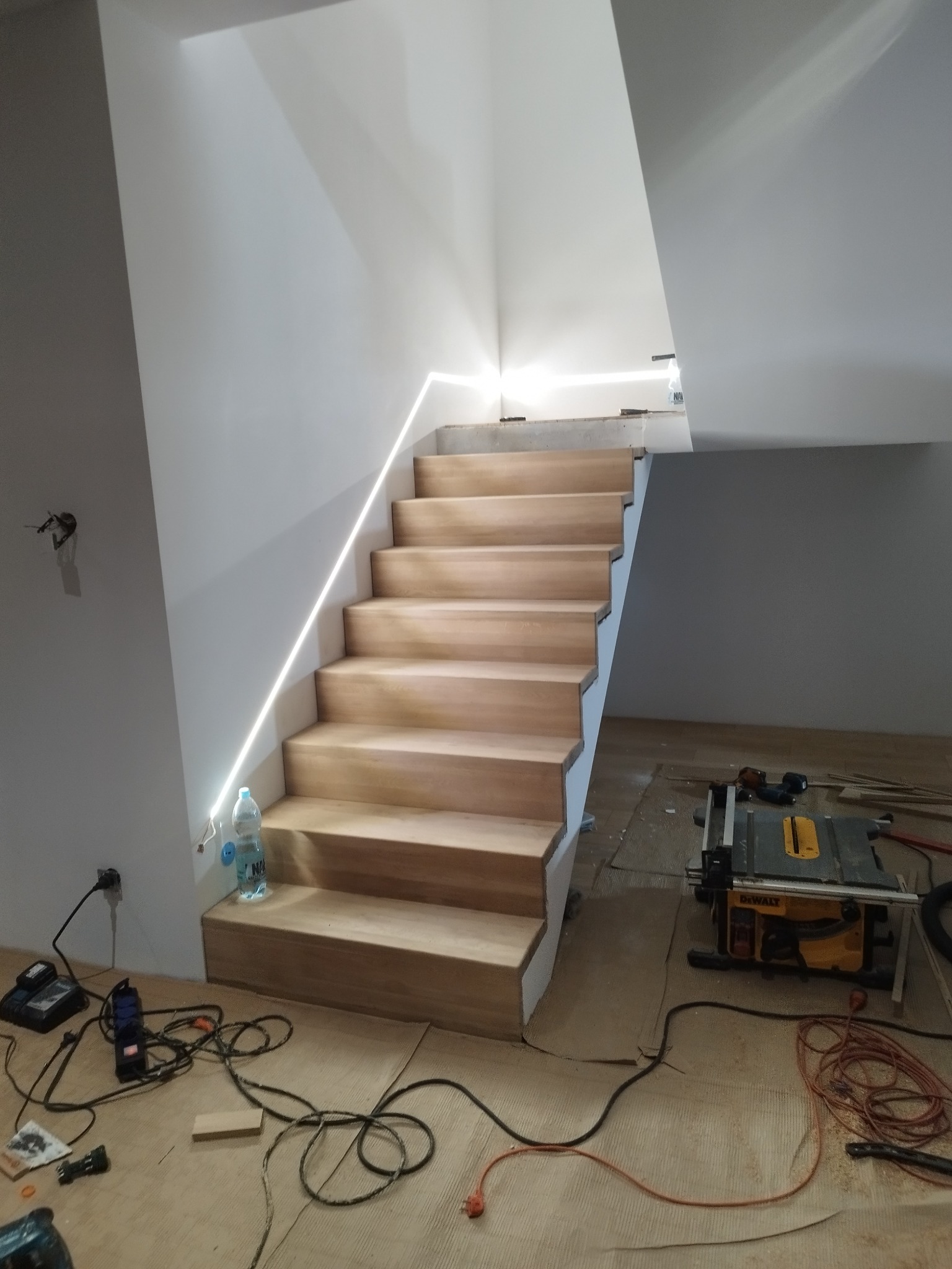 Jasne, drewniane schody z podświetleniem LED na ścianie. W tle widoczny sprzęt budowlany i butelka wody. Ujęcie z dołu, podkreślające nowoczesny design schodów.