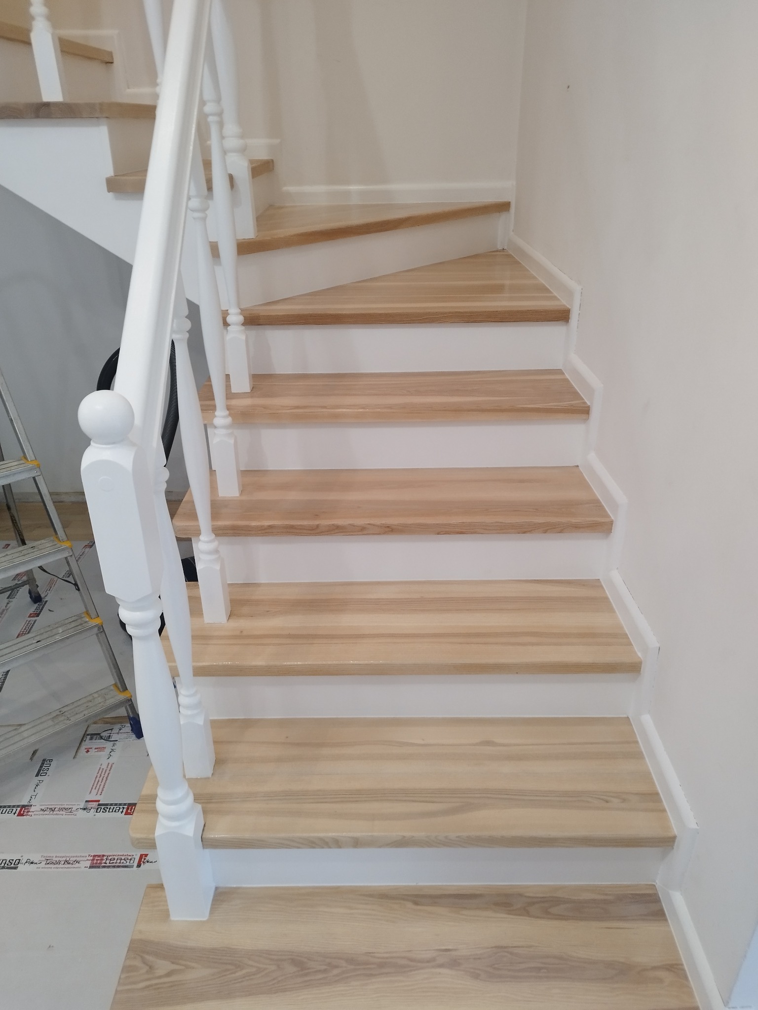 Jasne drewniane schody z białymi podstopnicami i balustradą, widok z dołu. Na podłodze folia ochronna. Detal wykończenia schodów.