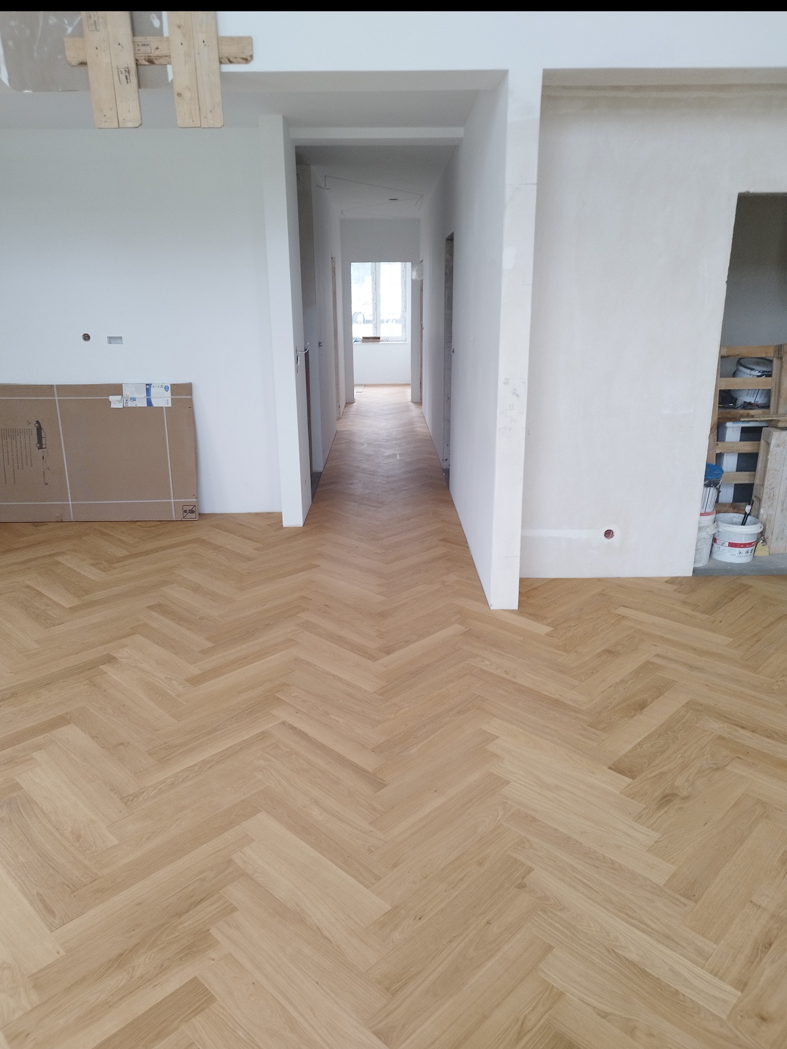 Jasny parkiet ułożony we wzór jodełki w długim korytarzu. Kartony przy ścianie sugerują etap wykończeniowy. Widok na dalszą część korytarza z oknem w tle.