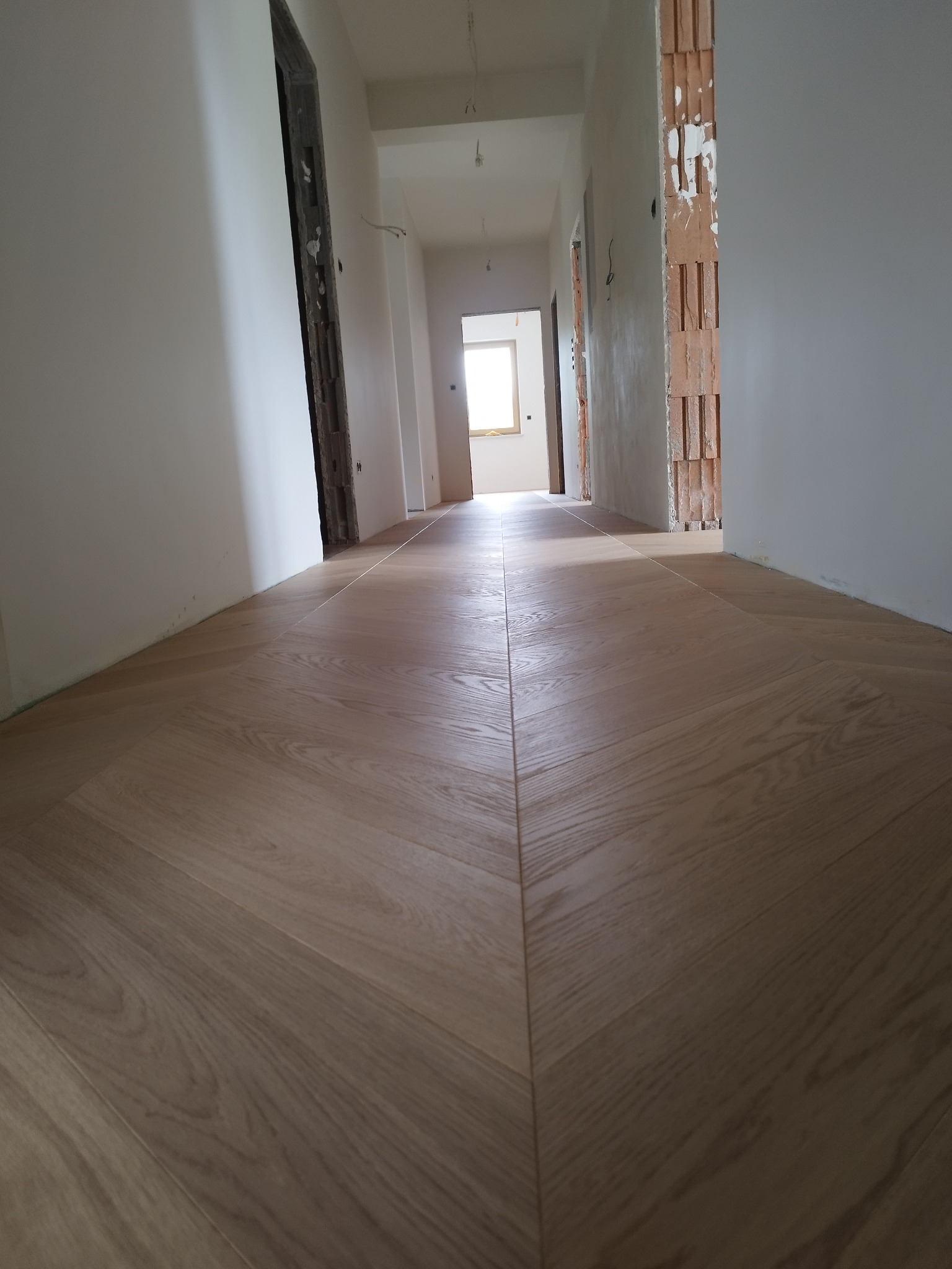 Ułożony parkiet w jodełkę francuską w korytarzu w trakcie remontu. Widoczne niedokończone ściany i futryny. Jasne drewno, naturalne światło z okna w głębi.