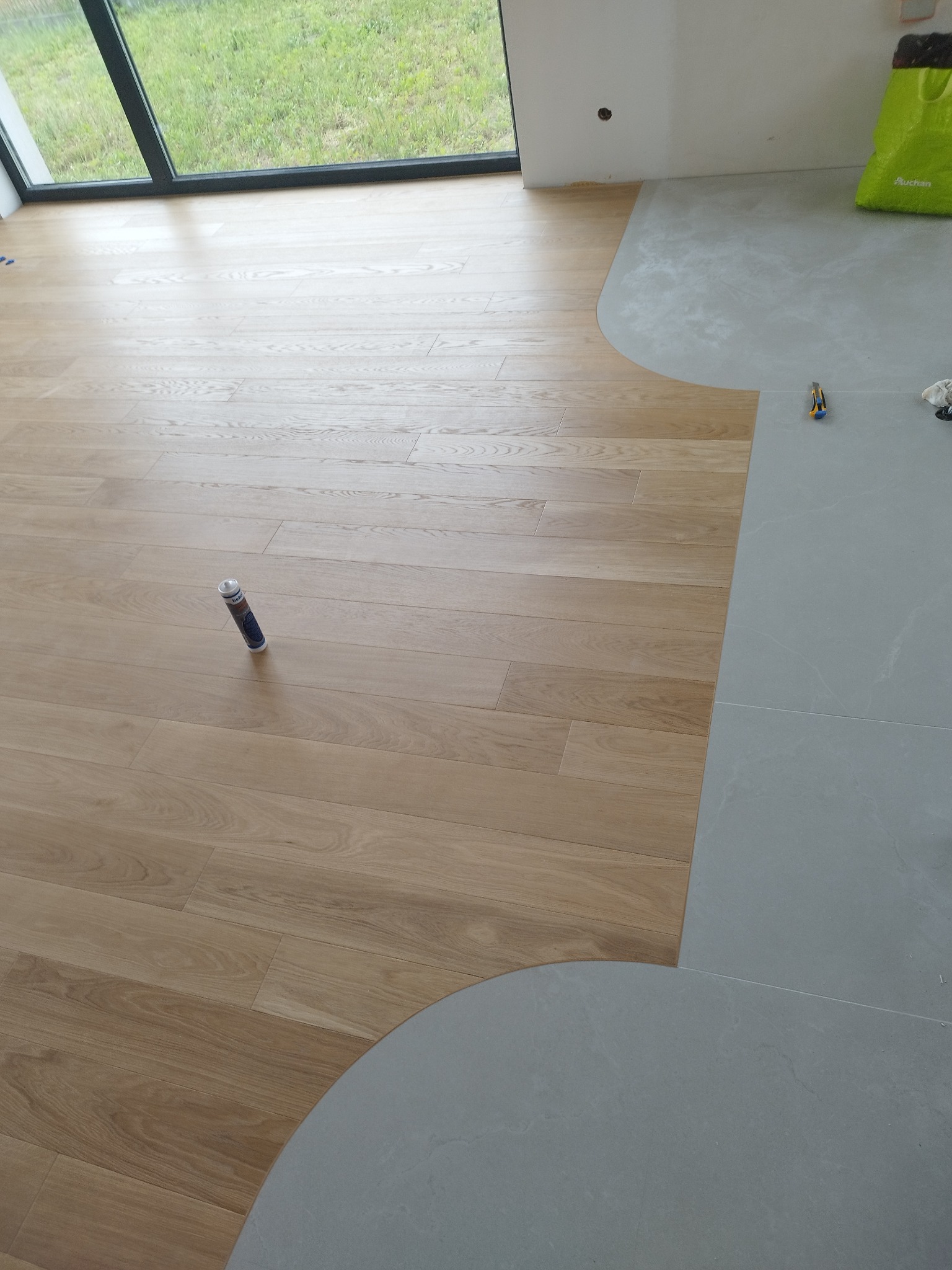 Połączenie paneli podłogowych z szarą posadzką. Widoczna tuba kleju, nóż i torba na tle okna. Układanie paneli w trakcie prac wykończeniowych.