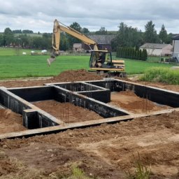 Maciej Kostuch - Fundamenty domu jednorodzinnego z izolacją, częściowo zasypane ziemią. W tle koparka i zabudowa wiejska. Ujęcie z poziomu gruntu, pochmurne niebo.