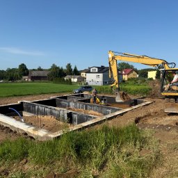 Maciej Kostuch - Fundament domu jednorodzinnego w trakcie budowy, z widocznym szalunkiem i pracującą koparką. W tle zabudowa wiejska i błękitne niebo.