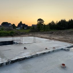 Maciej Kostuch - Zalana płyta fundamentowa z wystającymi prętami zbrojeniowymi na tle domów i zachodzącego słońca. Widoczny pracownik w ubraniu roboczym.