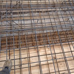 Maciej Kostuch - Zbrojenie fundamentów na drewnianym szalunku, widok z góry z fragmentem buta roboczego w rogu. Metalowe pręty tworzą siatkę. Etap przygotowawczy budowy.