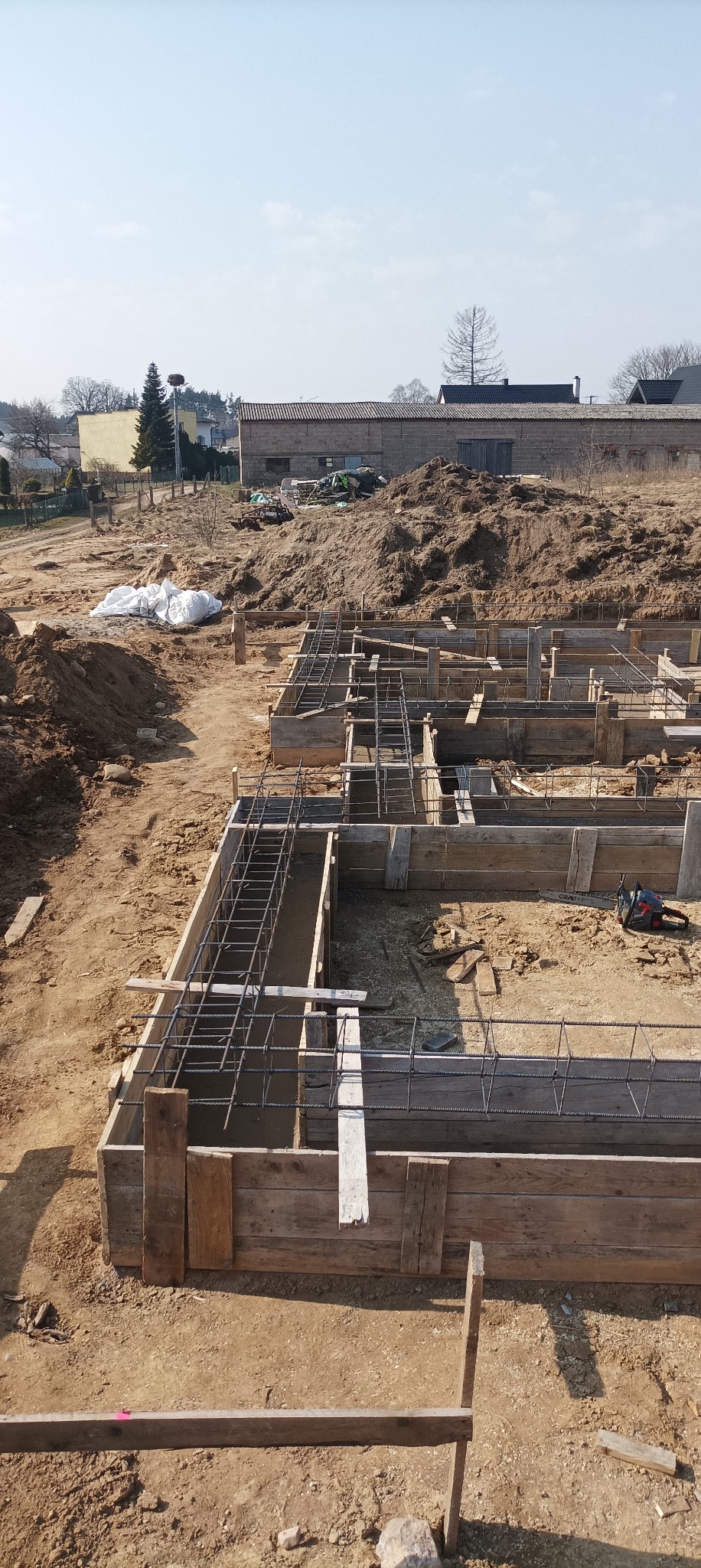 Fundamenty domu w trakcie budowy: drewniane szalunki zbrojone stalą, przygotowane do zalania betonem. W tle sterta ziemi i budynek gospodarczy.
