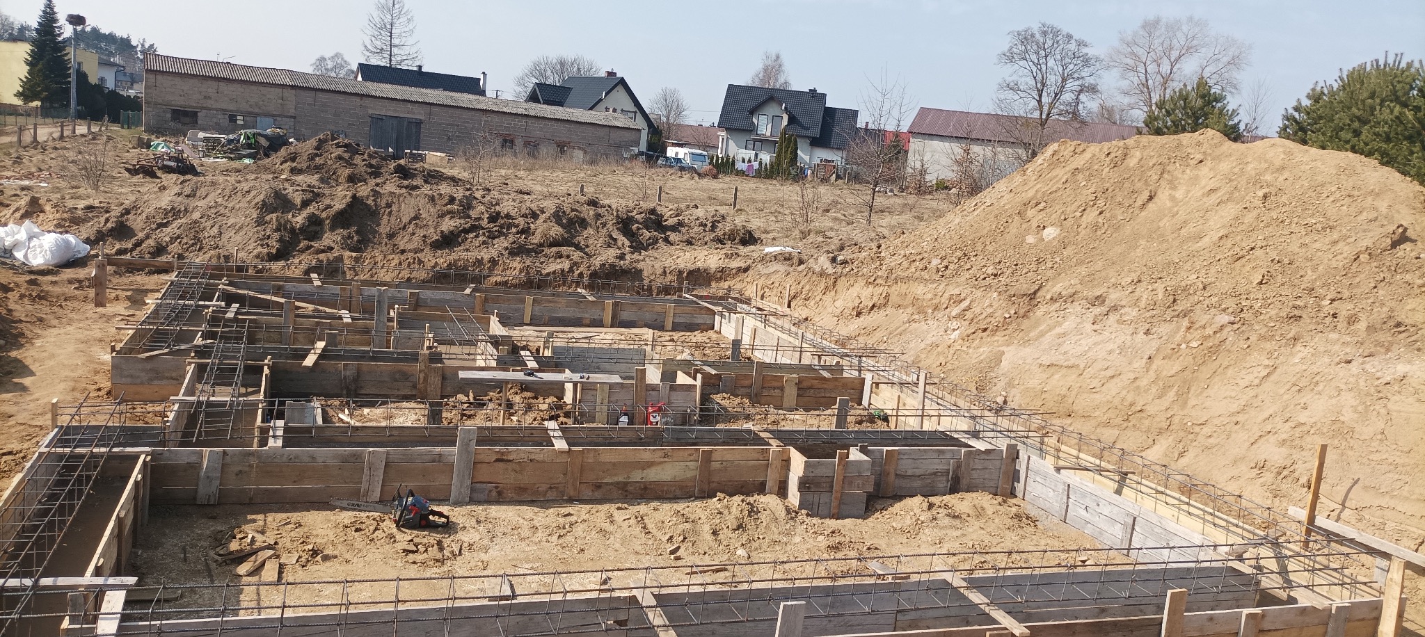 Szeroki kadr na szalunek fundamentów domu jednorodzinnego z widocznym zbrojeniem, ułożony na nierównym terenie z hałdami ziemi w tle.