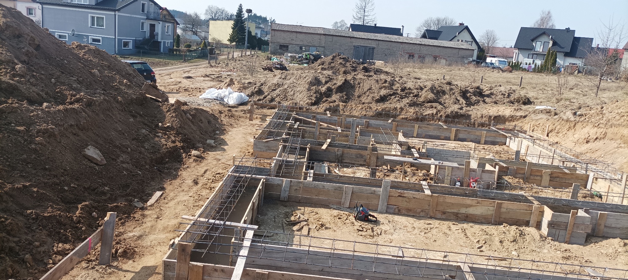 Ziemny wykop pod fundamenty domu z drewnianym szalunkiem i zbrojeniem stalowym. W tle zabudowa wiejska i hałda ziemi.