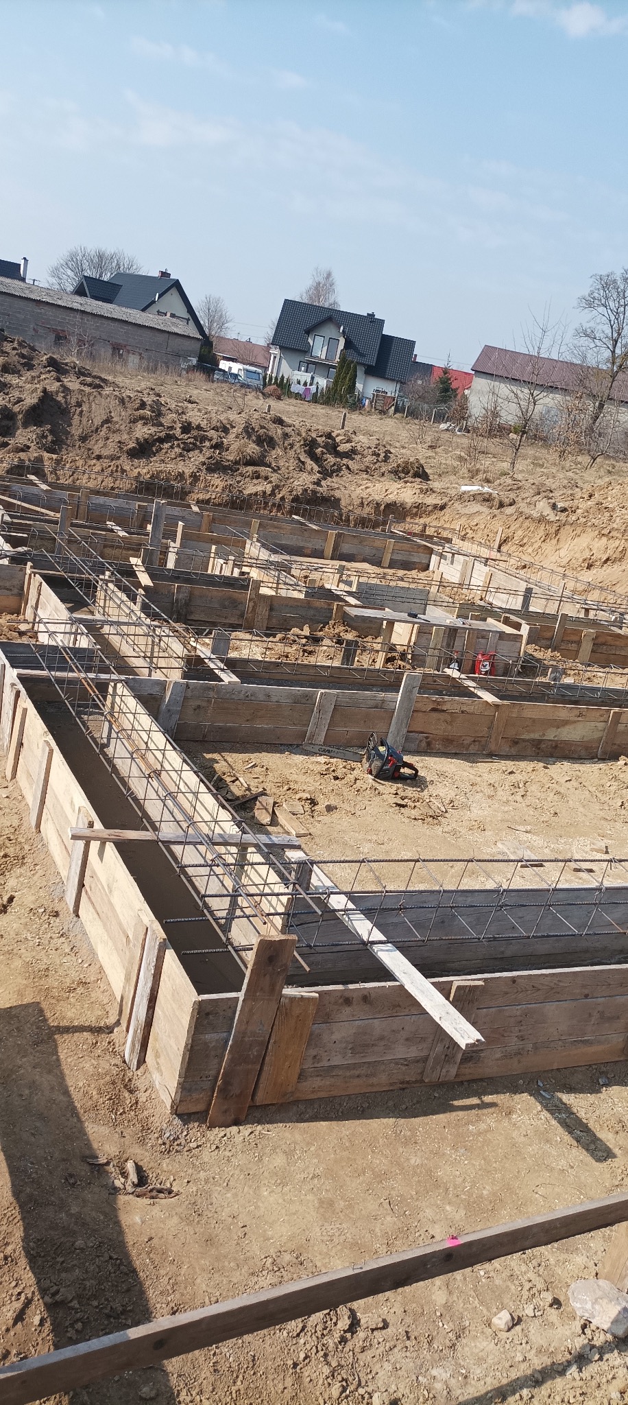Zbrojenie i szalunek fundamentów domu jednorodzinnego w słoneczny dzień. Widoczne drewniane formy i stalowe pręty zbrojeniowe. W tle zabudowa mieszkalna.