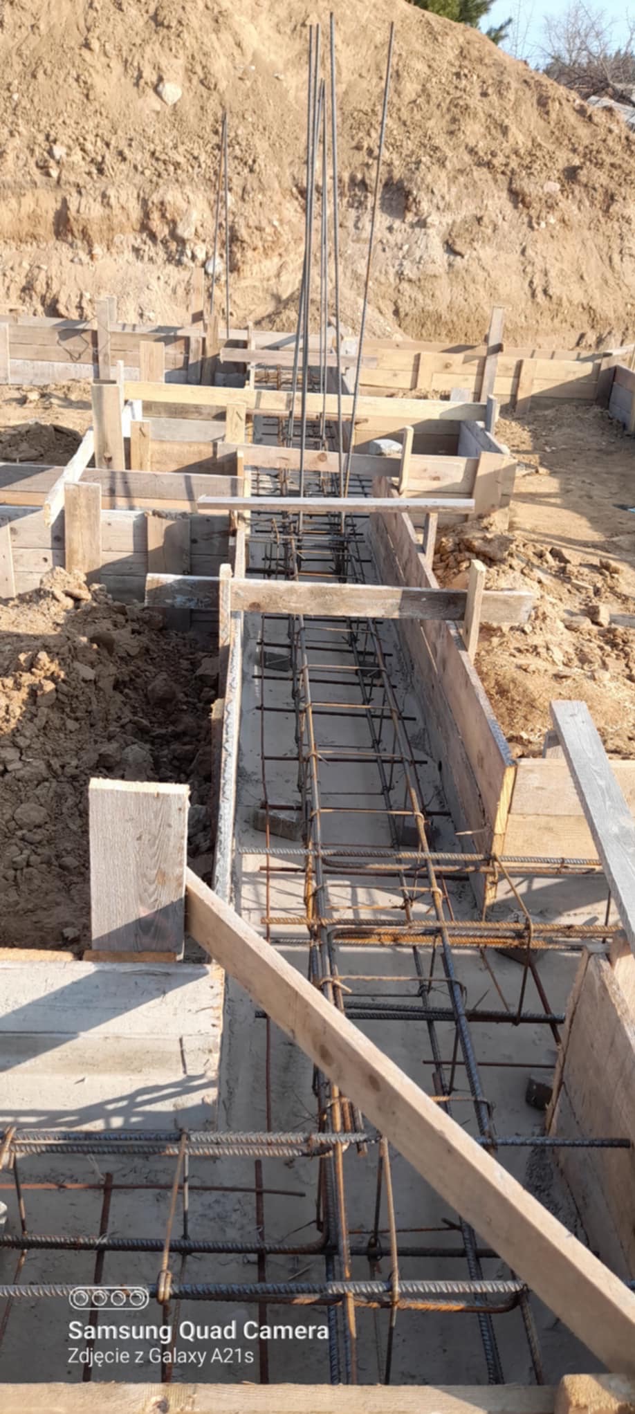 Zbrojenie fundamentów w drewnianym szalunku, widok z góry. Stalowe pręty tworzą siatkę w betonowych korytach. W tle ziemia i drewniane elementy konstrukcyjne.
