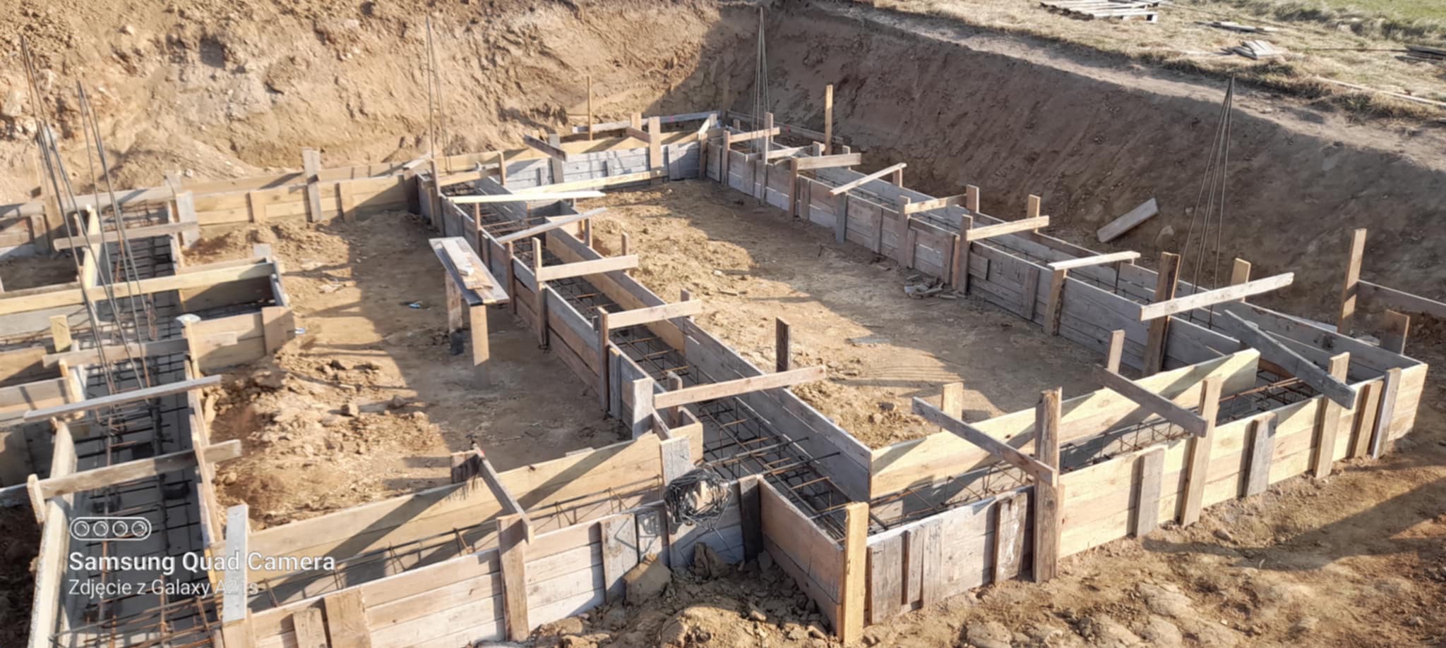 Szeroki kadr na wykop z drewnianym szalunkiem i zbrojeniem fundamentów domu jednorodzinnego. Widok z góry, ziemisty teren.