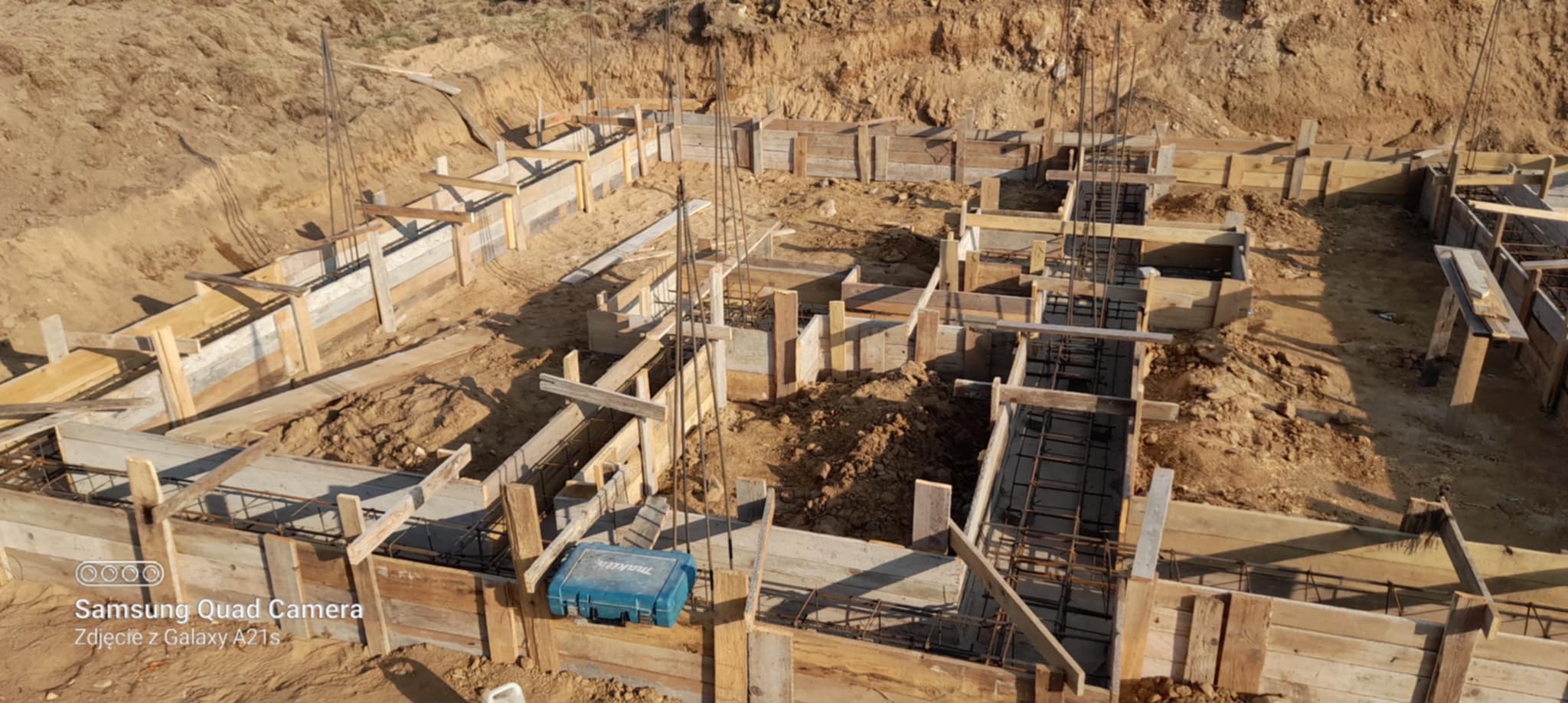 Zbrojenie i szalunek fundamentów domu jednorodzinnego. Widoczne pręty zbrojeniowe i drewniane deski szalunkowe. Na pierwszym planie niebieska skrzynka narzędziowa.
