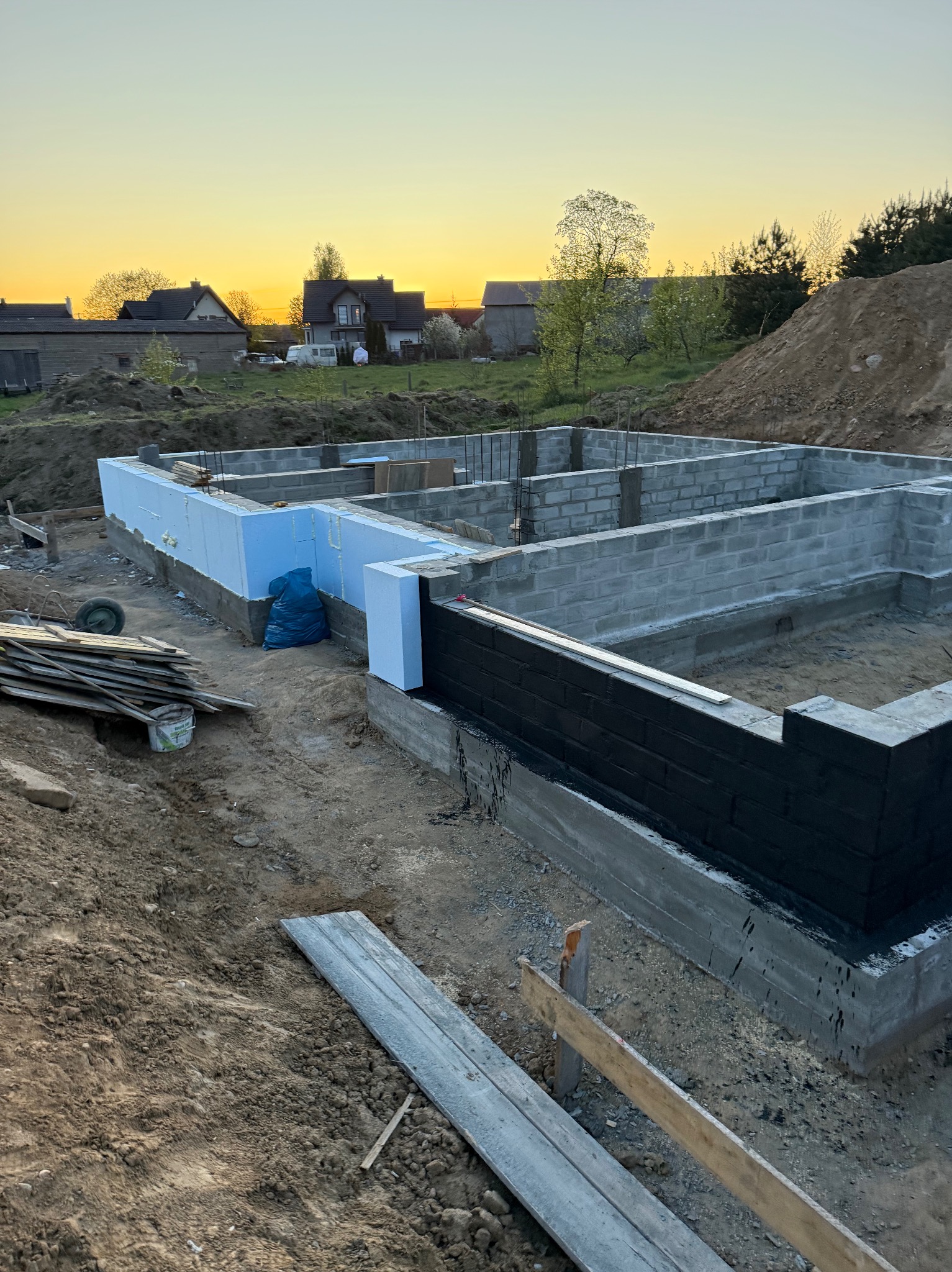 Fundamenty domu w trakcie izolacji: czarna hydroizolacja i niebieski styropian. Widoczne bloczki betonowe i zbrojenie. W tle zabudowa jednorodzinna o zmierzchu.