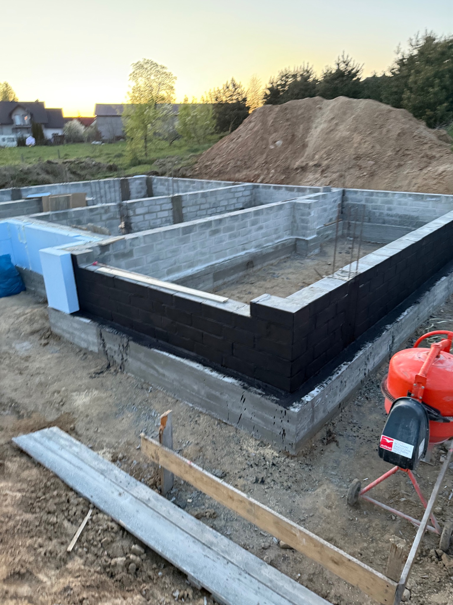 Fundamenty domu w trakcie budowy: bloczki betonowe, izolacja pionowa pomalowana na czarno, styropian, widoczna betoniarka. W tle hałda ziemi i zabudowa jednorodzinna.