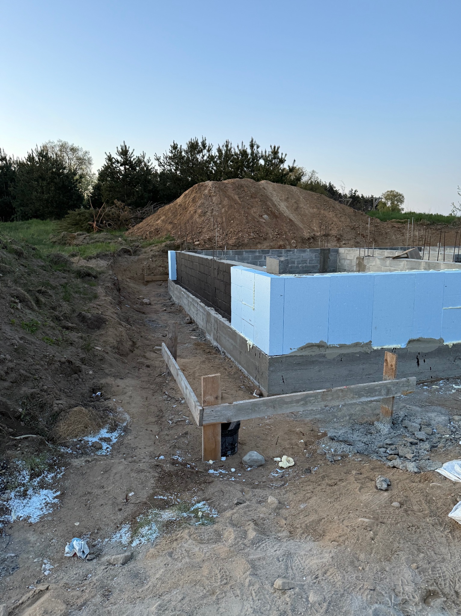Fundamenty domu w trakcie izolacji, z widoczną niebieską warstwą styropianu i betonową konstrukcją. W tle usypana ziemia i drzewa.