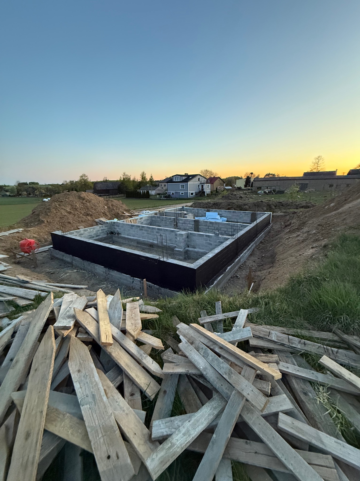 Fundamenty domu jednorodzinnego z izolacją, widok z góry na stos desek, w tle domy i zachodzące słońce.