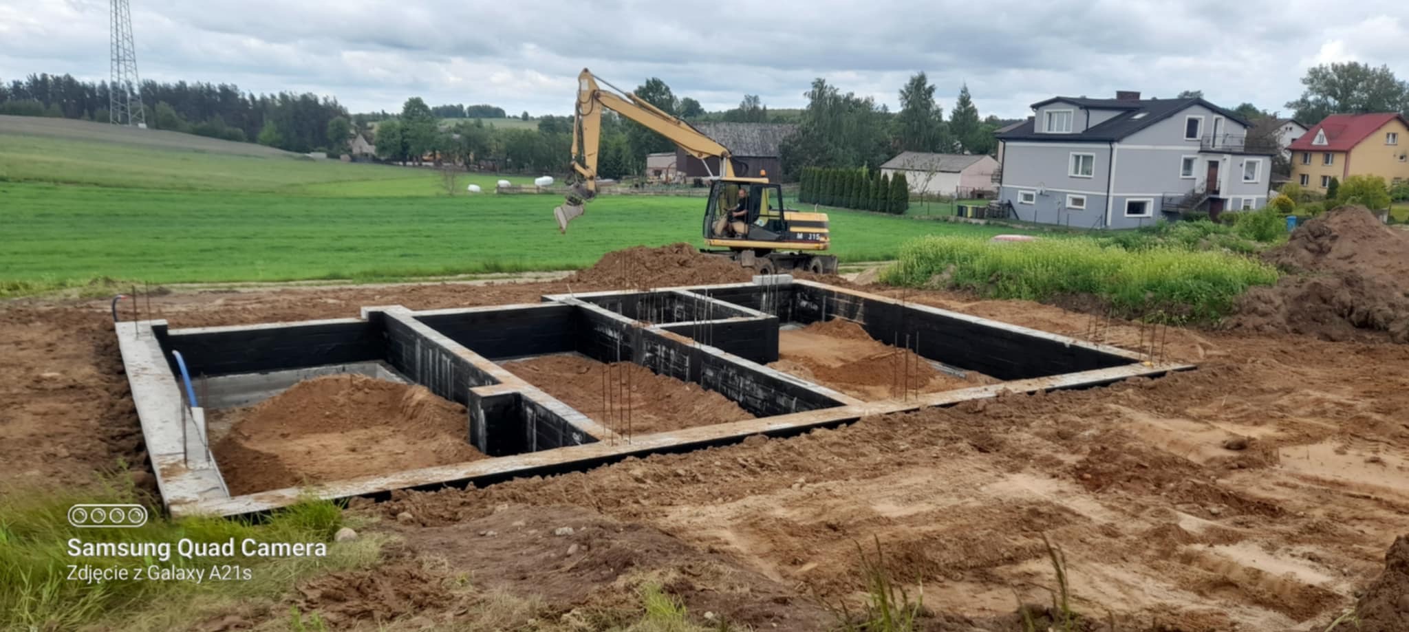 Fundamenty domu jednorodzinnego z izolacją, częściowo zasypane ziemią. W tle koparka i zabudowa wiejska. Ujęcie z poziomu gruntu, pochmurne niebo.