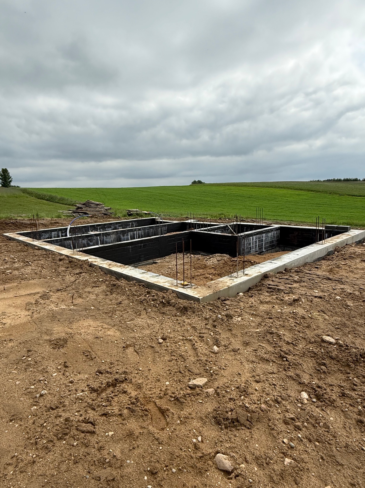 Fundament domu jednorodzinnego zbrojony stalą, zabezpieczony izolacją pionową, Kamienica Szlachecka. Widok na zielone pole i pochmurne niebo.