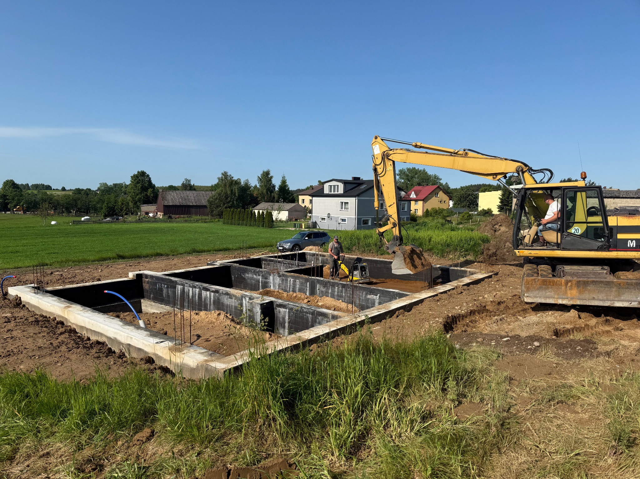 Fundament domu jednorodzinnego w trakcie budowy, z widocznym szalunkiem i pracującą koparką. W tle zabudowa wiejska i błękitne niebo.