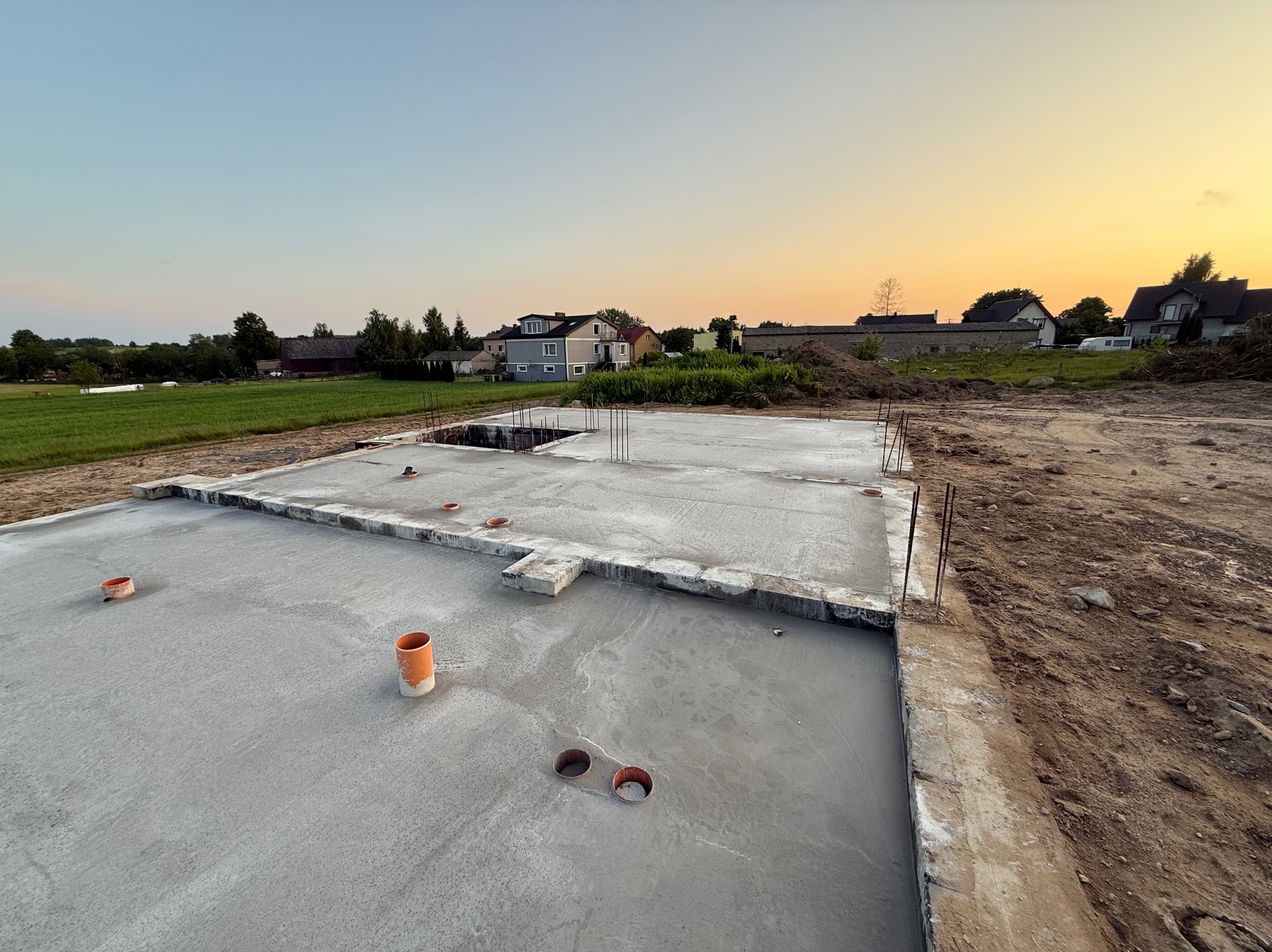 Wylana płyta fundamentowa z wystającymi rurami instalacyjnymi i prętami zbrojeniowymi na tle zabudowy jednorodzinnej o zmierzchu.